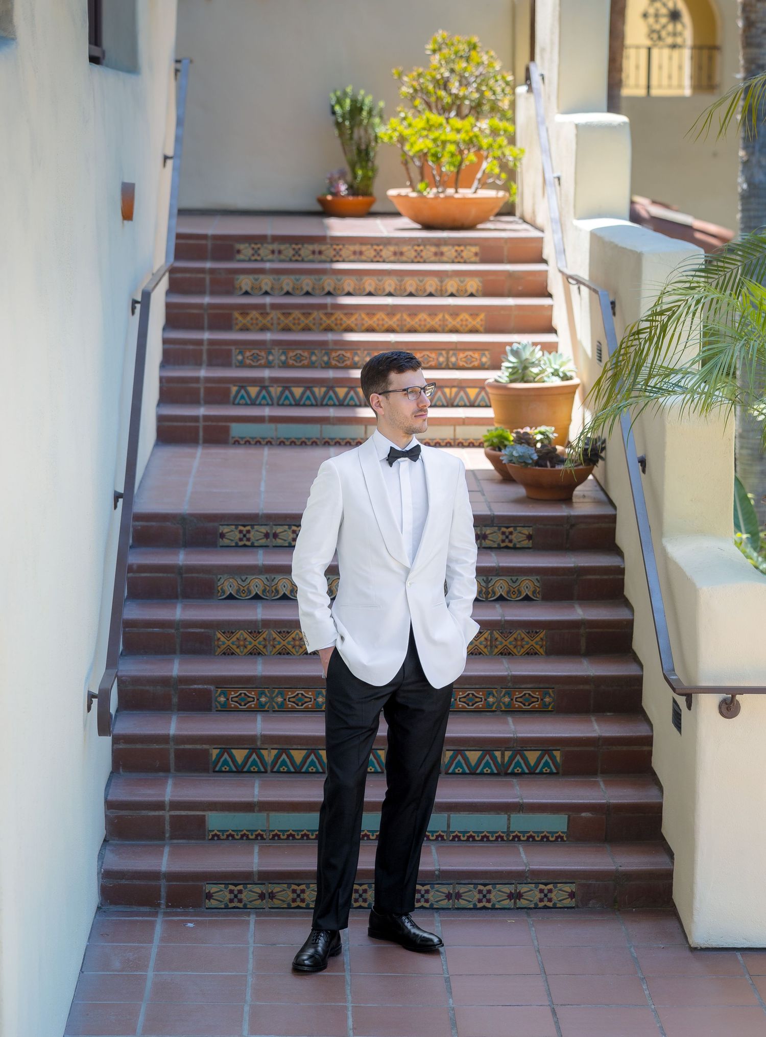 Groom in white tuxedo jacket and black bow tie stands on decorative tiled steps at an outdoor wedding venue.