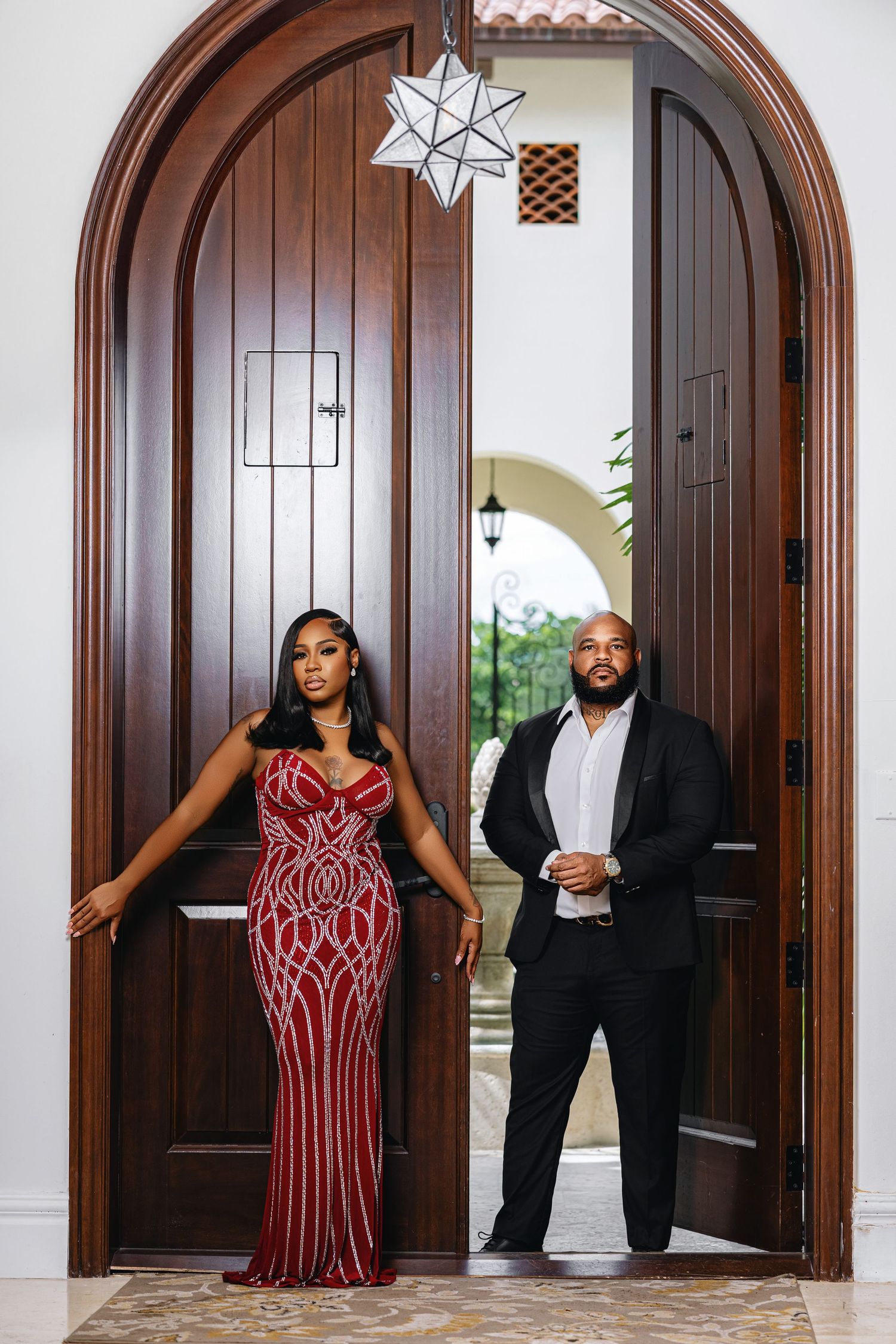 Elegant couple posing in grand arched wooden doorway, woman in red sequin gown, man in black suit.