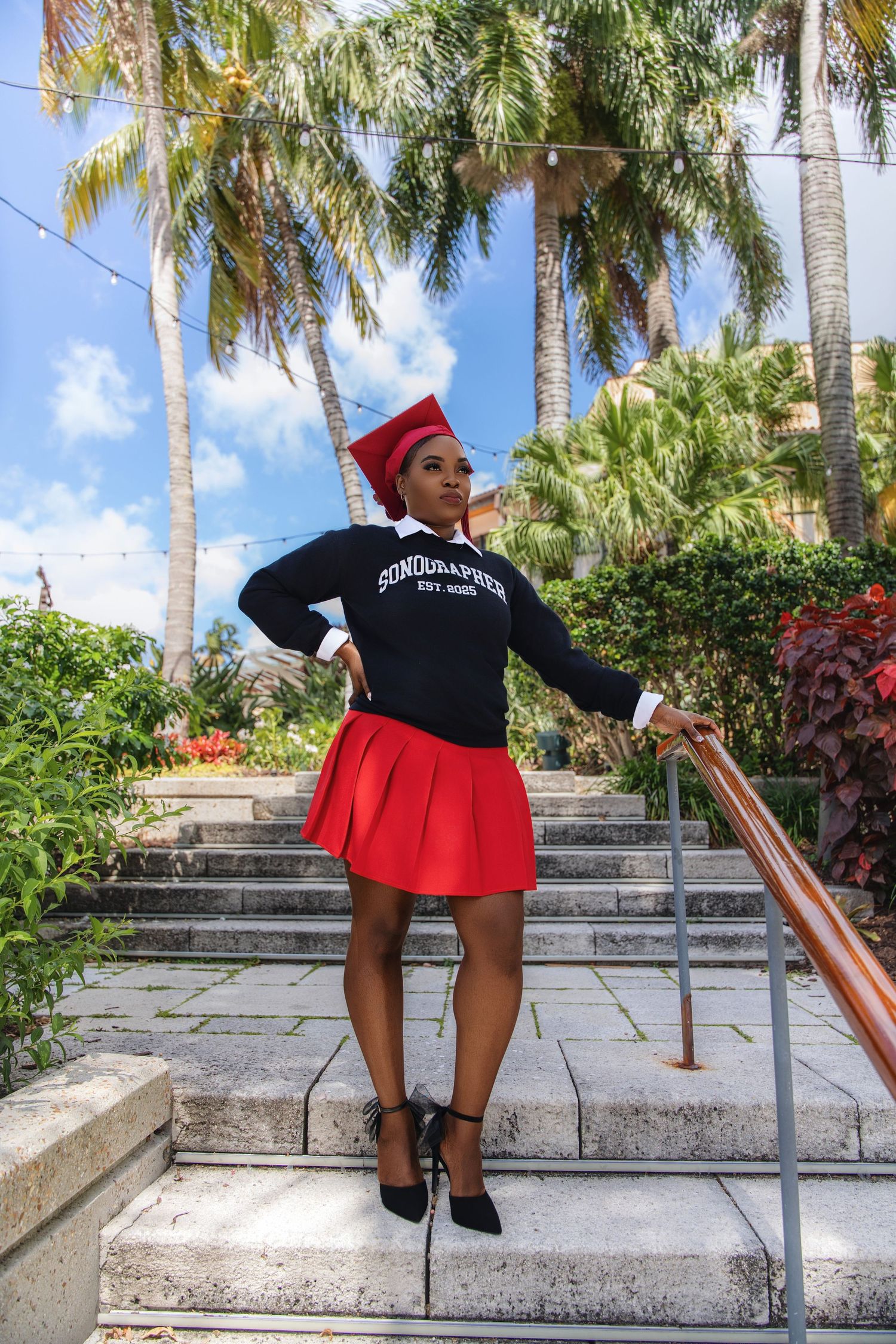 Young Black woman in red graduation cap, navy Sonographer sweatshirt, and red skirt poses on outdoor steps with palm trees.