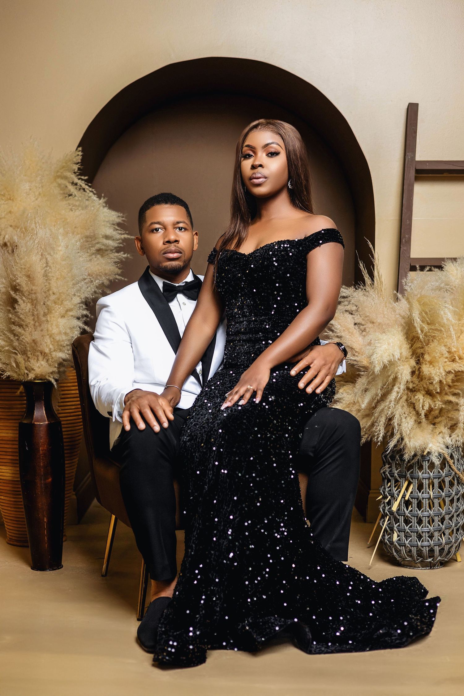 Couple in formal attire posing, man seated in white tuxedo, woman standing in black sequin mermaid gown.