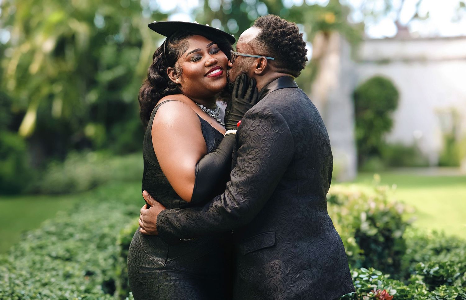 A joyful couple embraces and shares a kiss outdoors surrounded by lush green gardens.