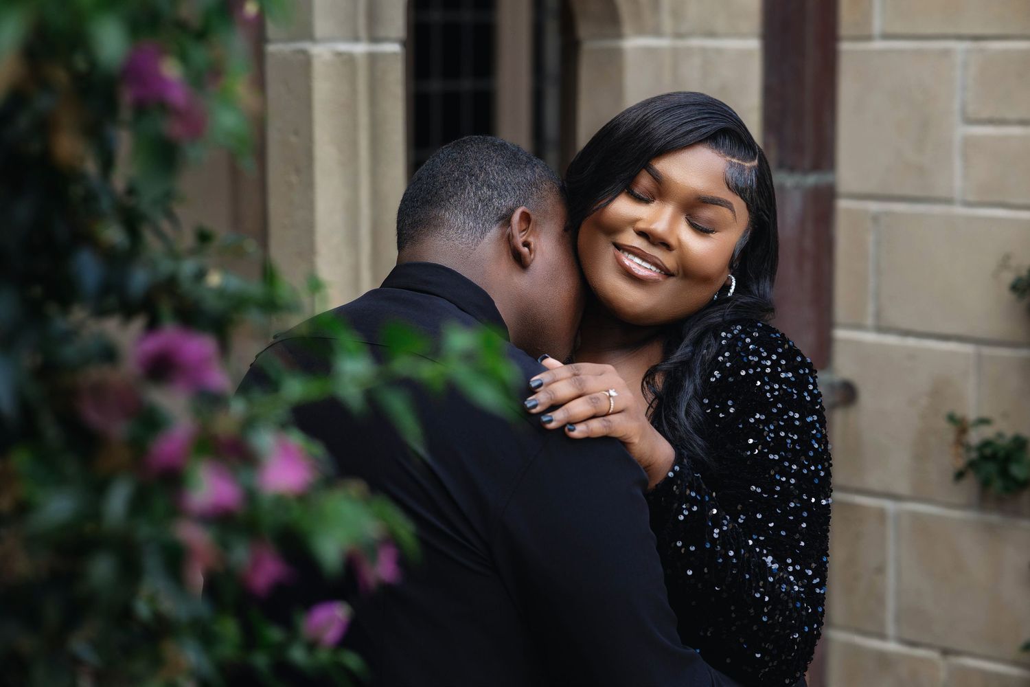 Joyful woman with engagement ring hugging her partner near purple flowers and stone building exterior.