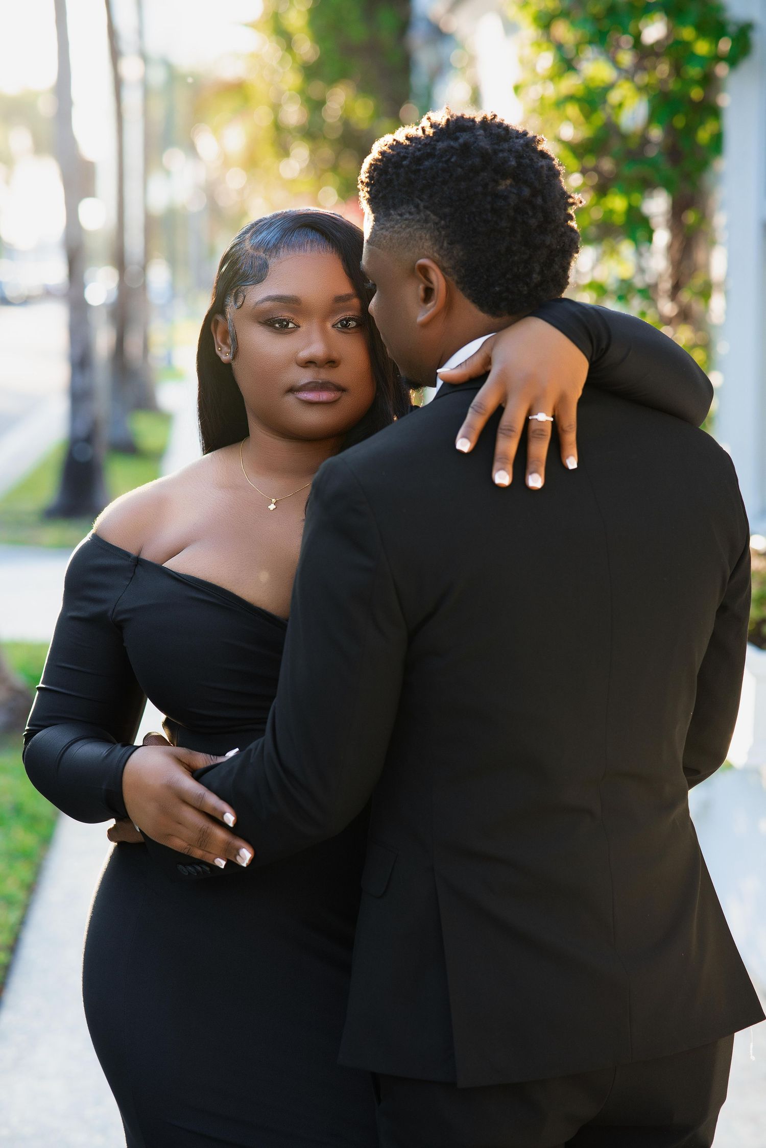 Couple in black formal wear sharing an intimate embrace outdoors in sunlight.