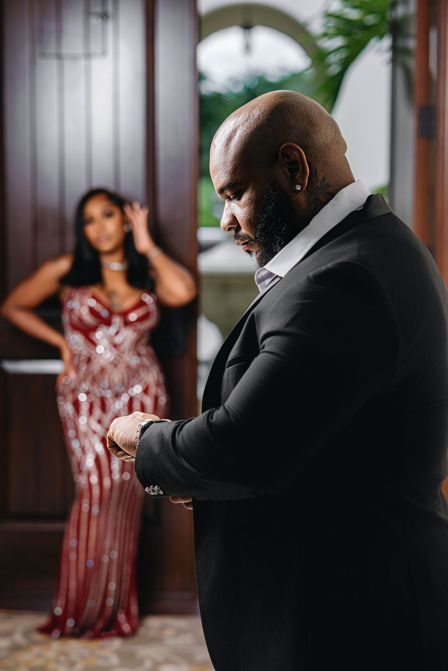 Dapper man in black suit adjusting cufflinks with woman in red sequin dress in background.