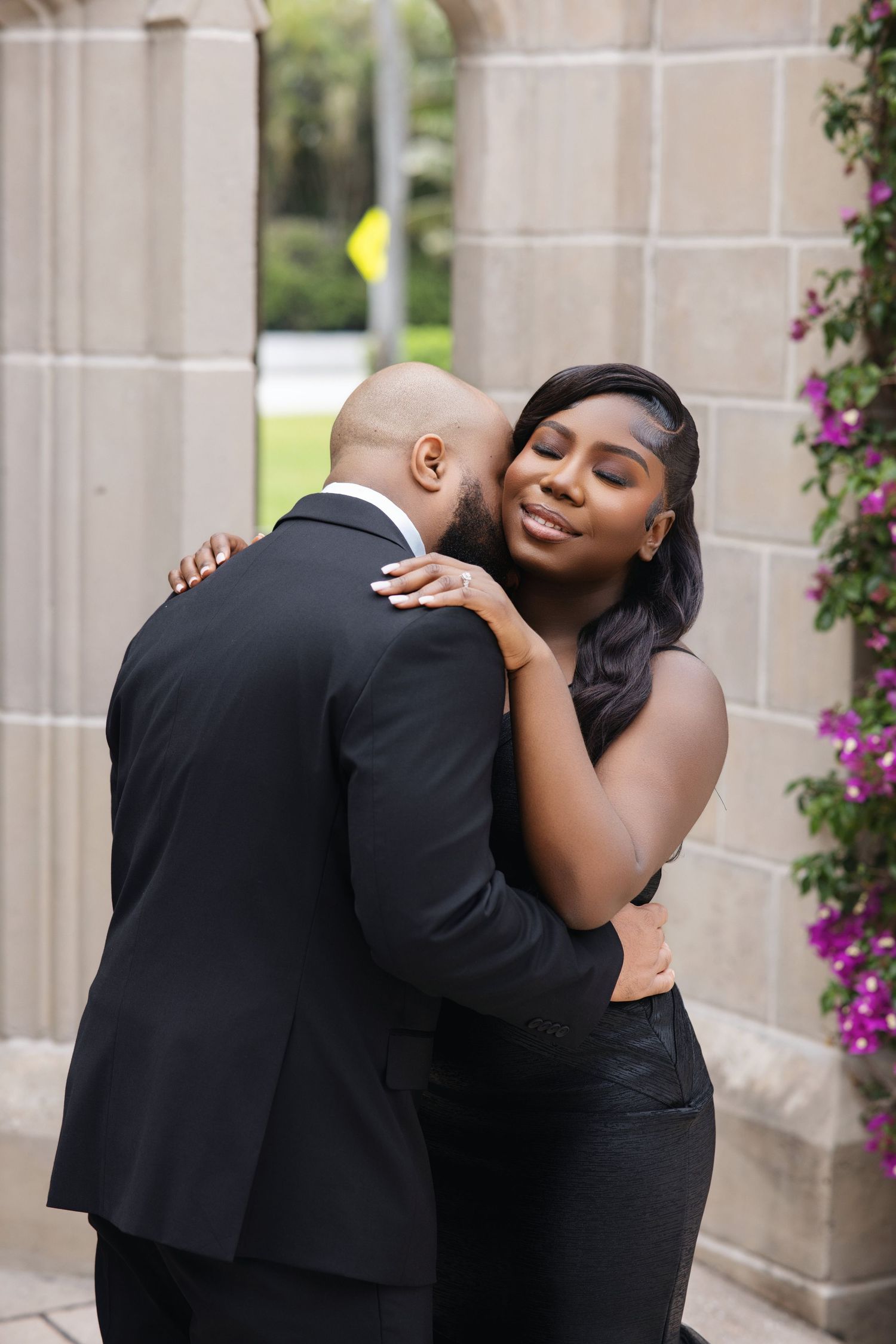 Black couple sharing a tender embrace near stone columns adorned with purple bougainvillea.