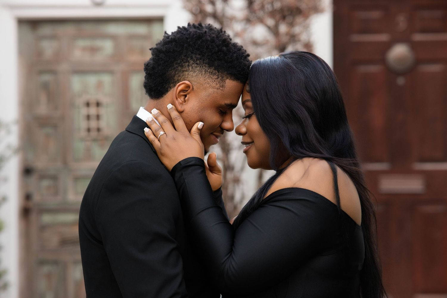 An elegant couple dressed in matching black outfits touch foreheads in a romantic pose against a rustic backdrop.