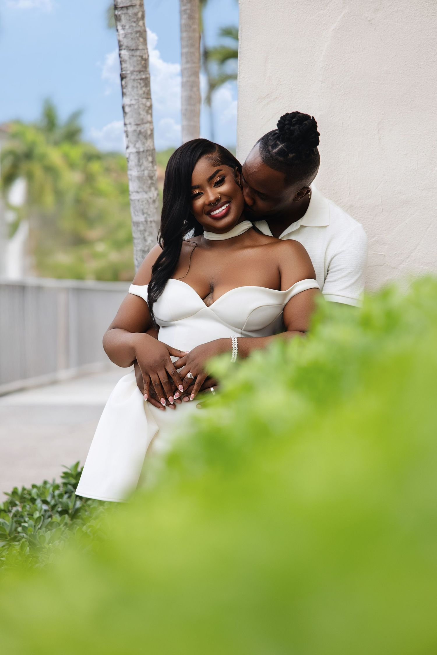 A Black couple poses romantically against a white wall, with the man kissing the woman's cheek.