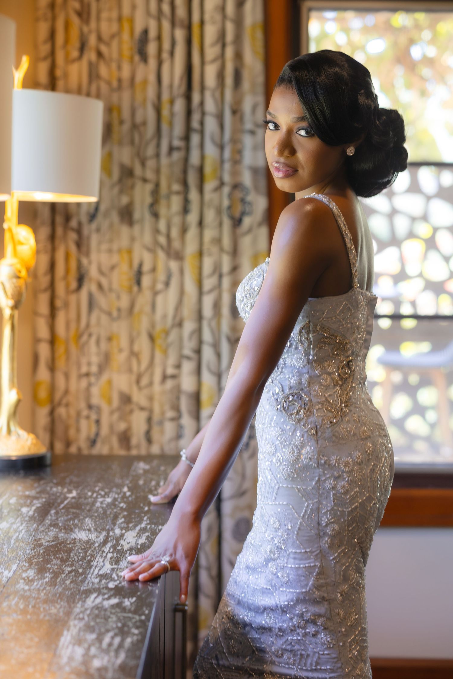 Elegant bride in a fitted white lace gown poses gracefully in a warmly lit room with floral curtains.