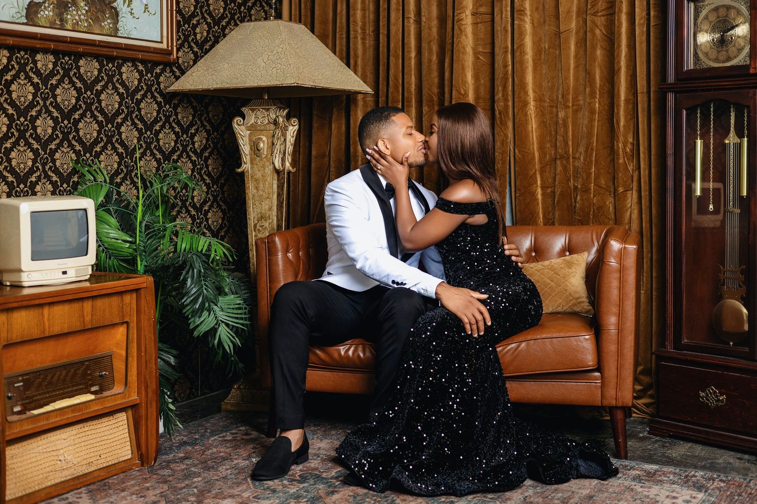 Couple sharing a kiss on leather sofa in vintage-styled room with retro decor and patterned wallpaper.