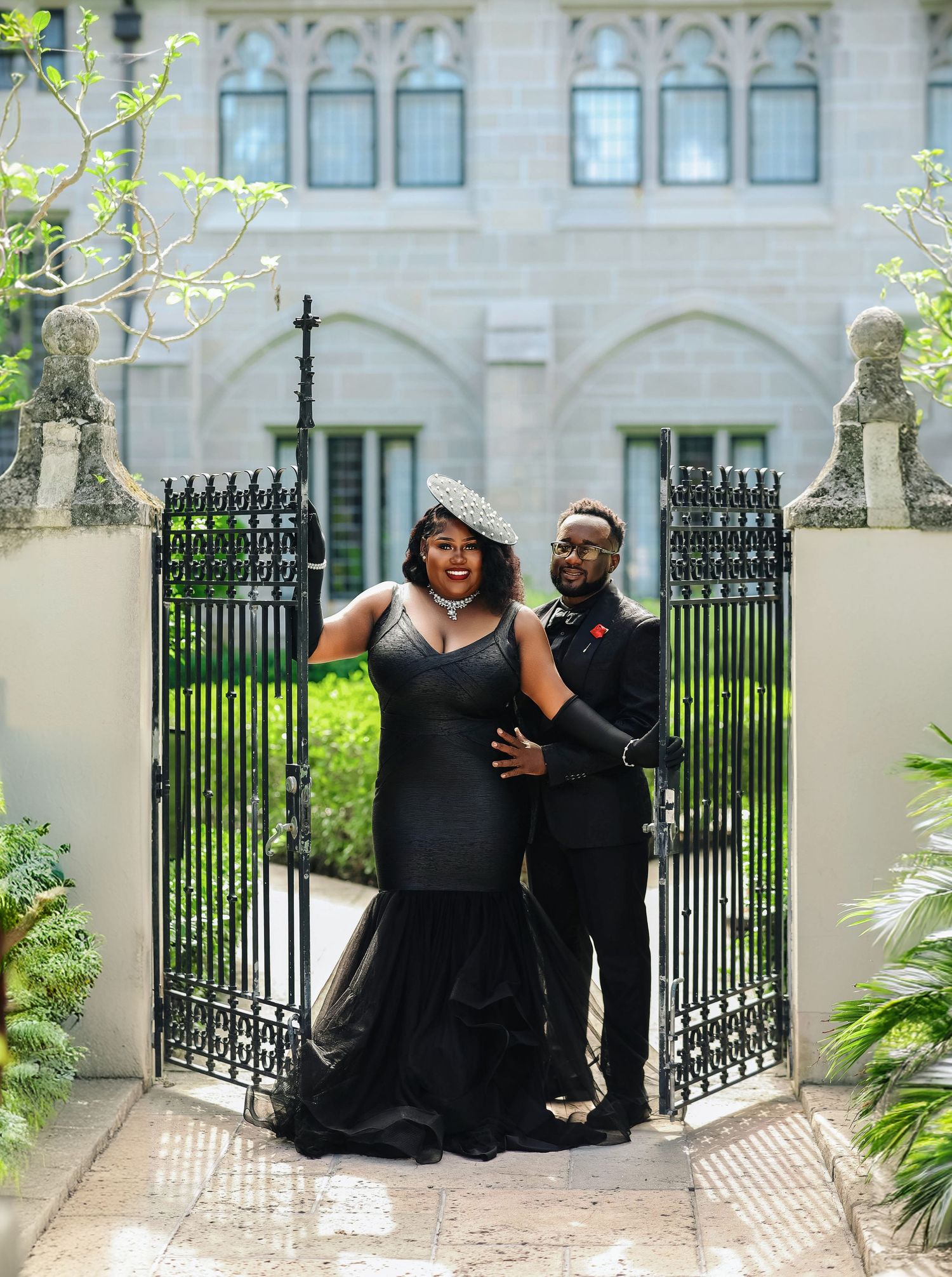 A couple in elegant black formal attire pose confidently before ornate iron gates of a historic estate.
