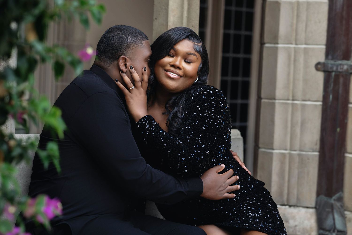 Happy couple sitting together outdoors, woman smiling while embracing man near flowering stone building.