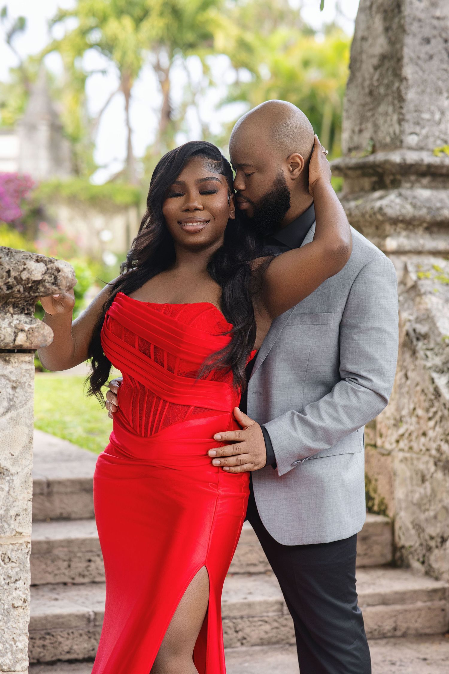 Romantic couple posing outdoors, woman in red mermaid gown, man in gray suit kissing her cheek.