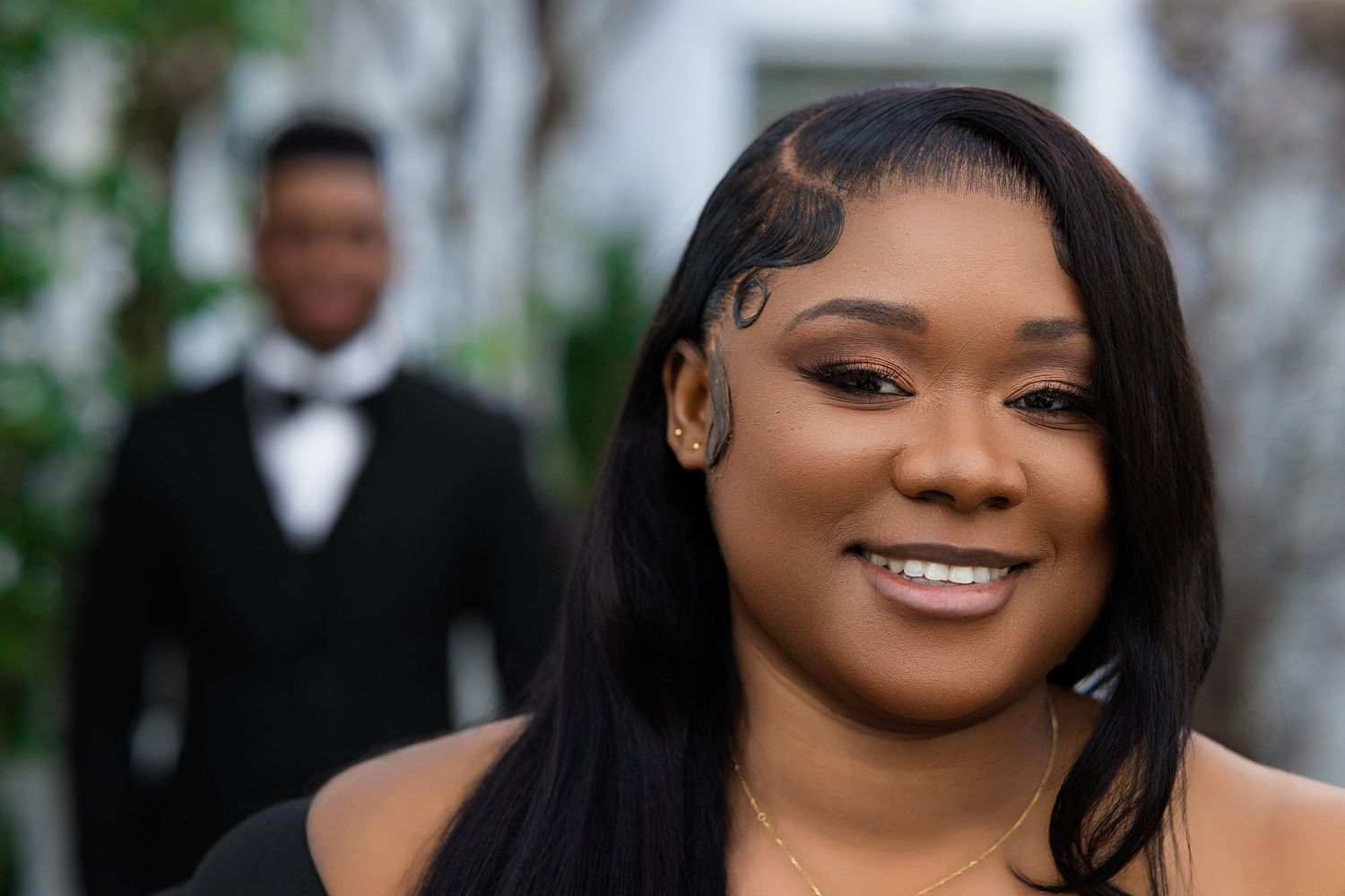 Close-up portrait of smiling woman with sleek hair, man in tuxedo blurred behind.