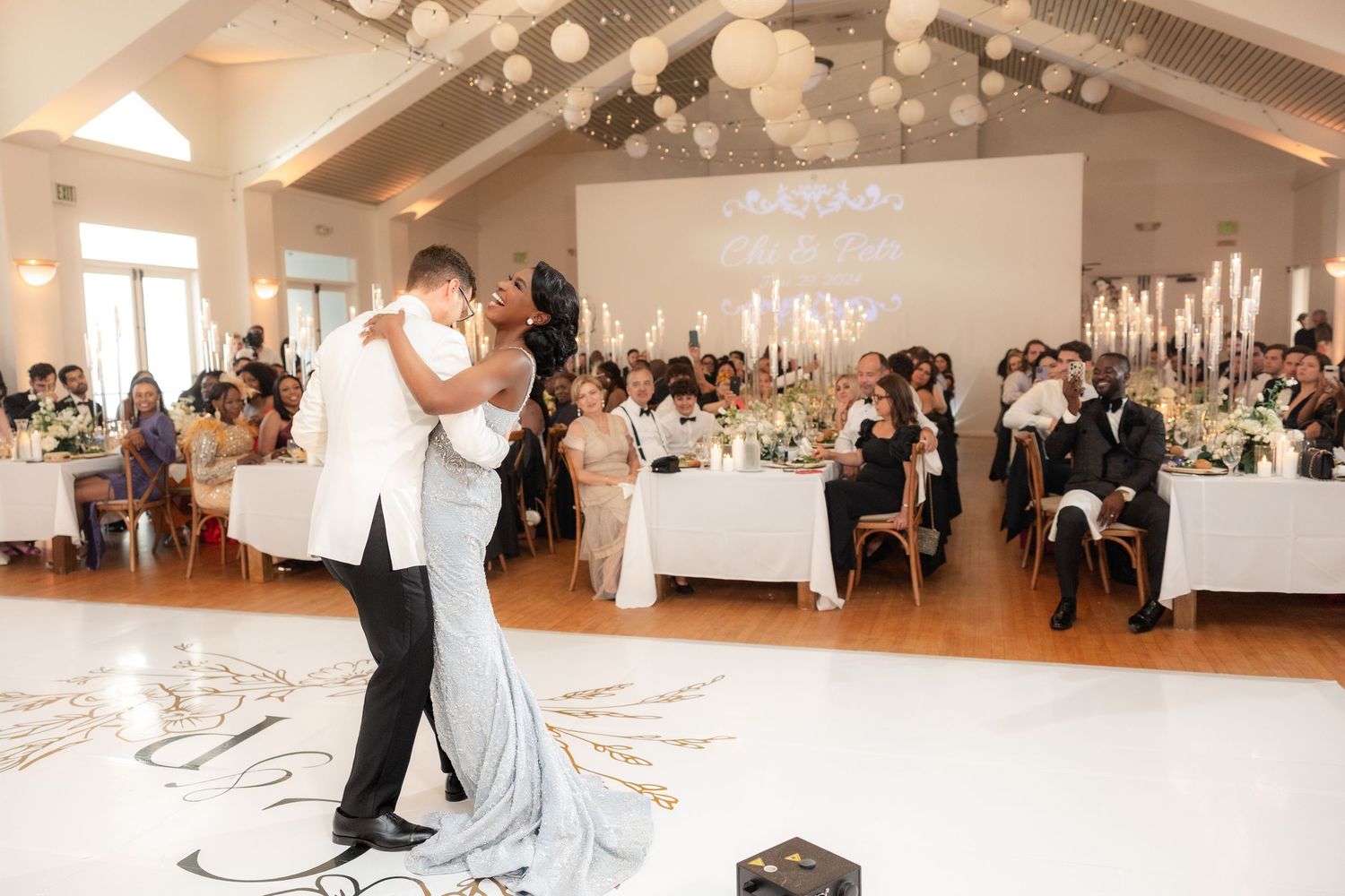 Bride and groom share first dance at elegant wedding reception with white decor, chandeliers, and guests seated around the dance floor.