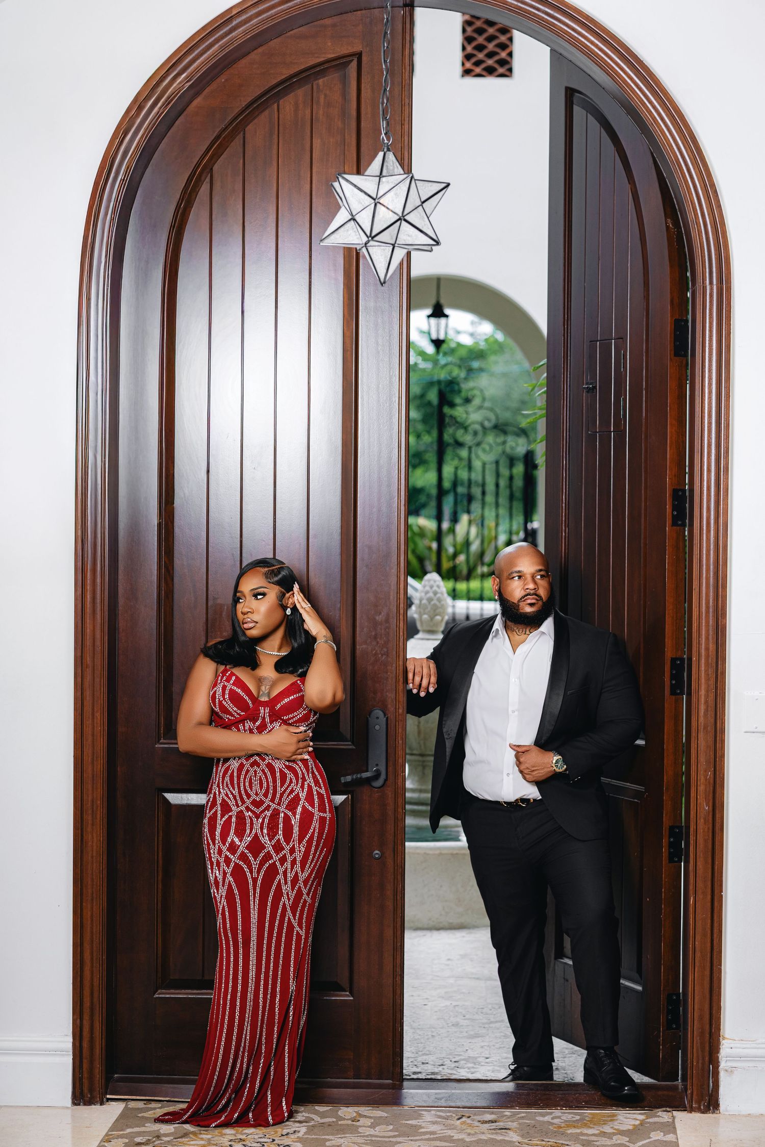 Couple posing in grand arched wooden doorway, woman in red sequined gown and man in black suit.