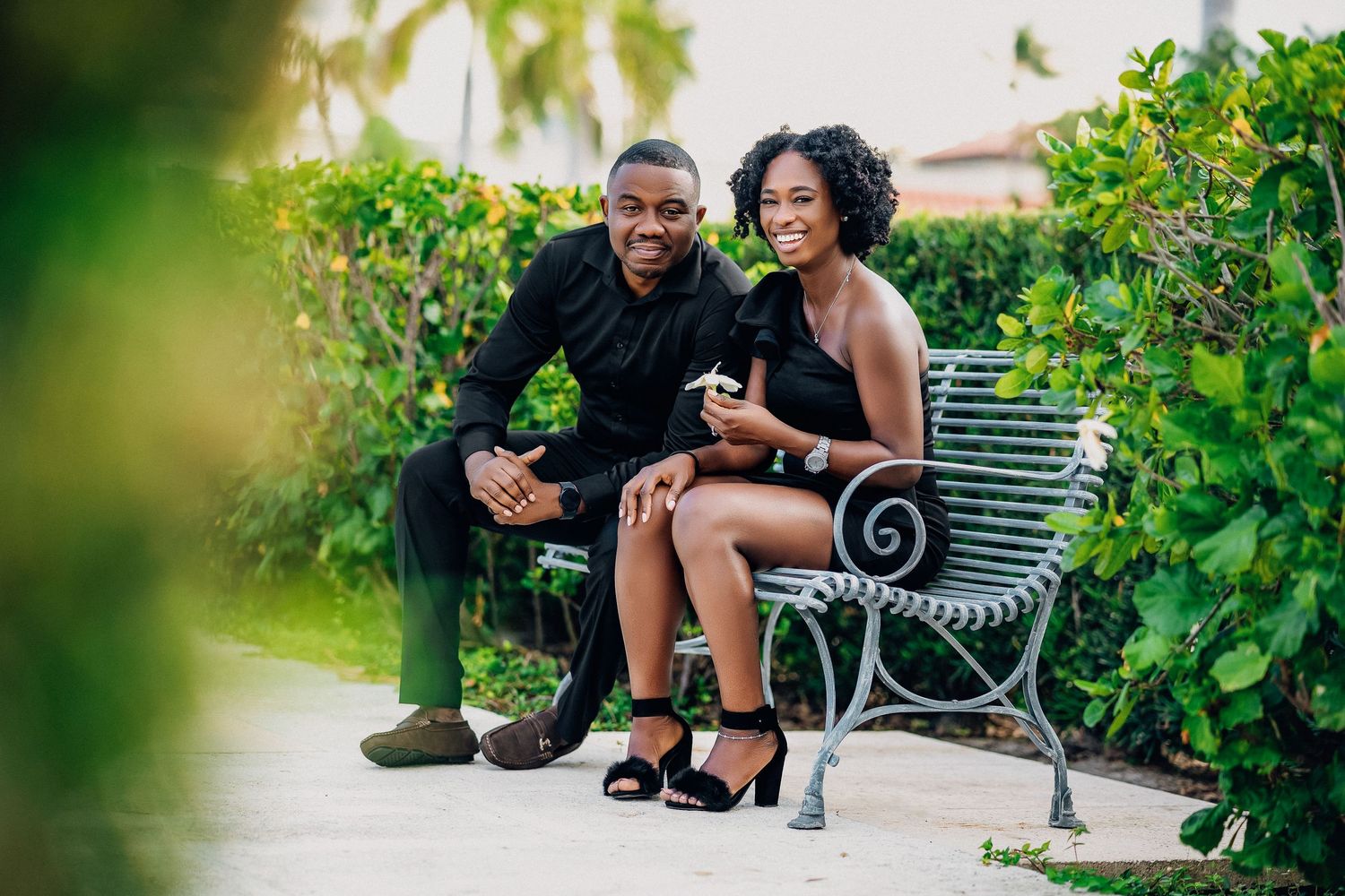 A smiling couple in black attire sit together on an ornate bench surrounded by tropical greenery.