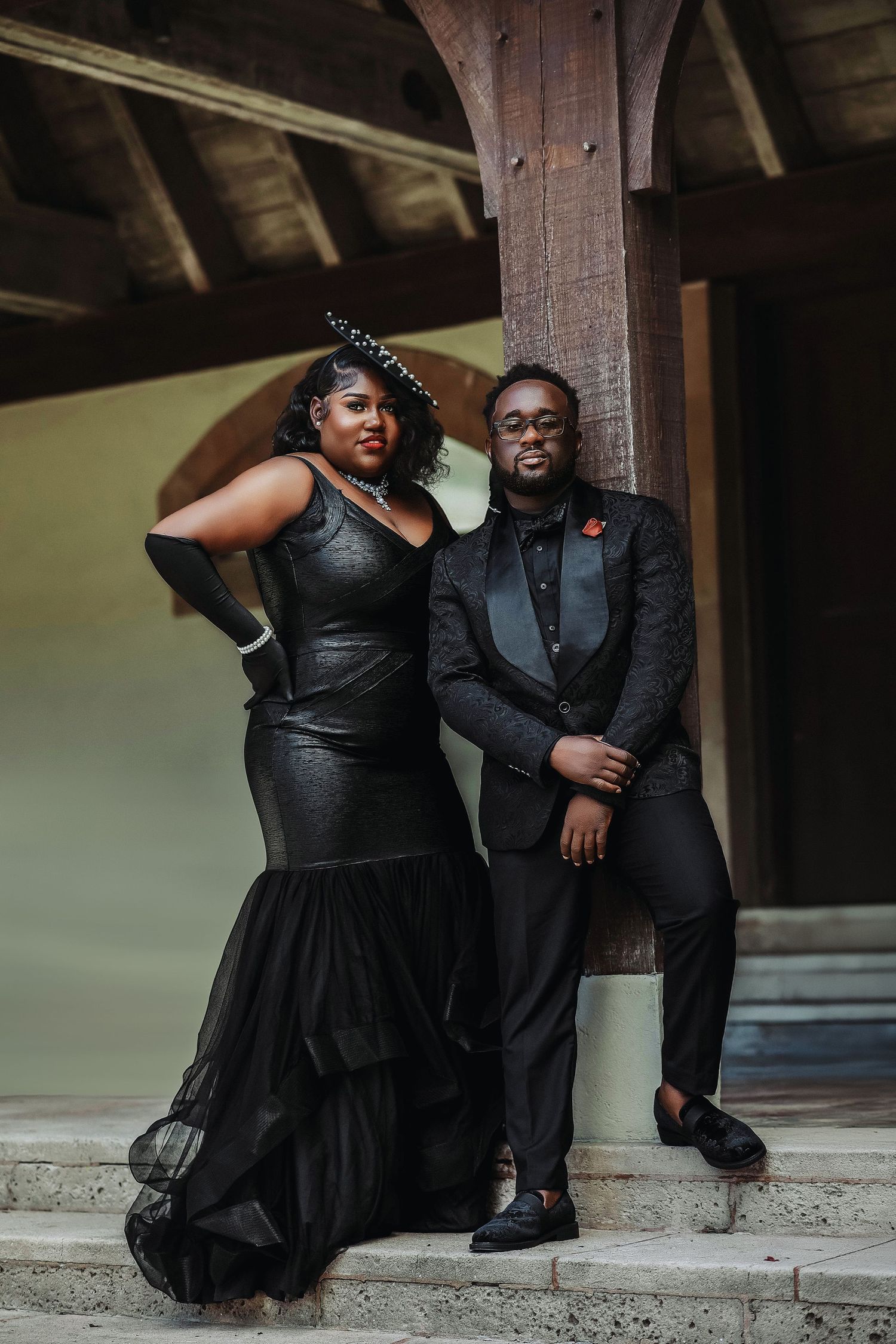 A couple in matching black formal attire pose on steps beneath a rustic wooden structure.