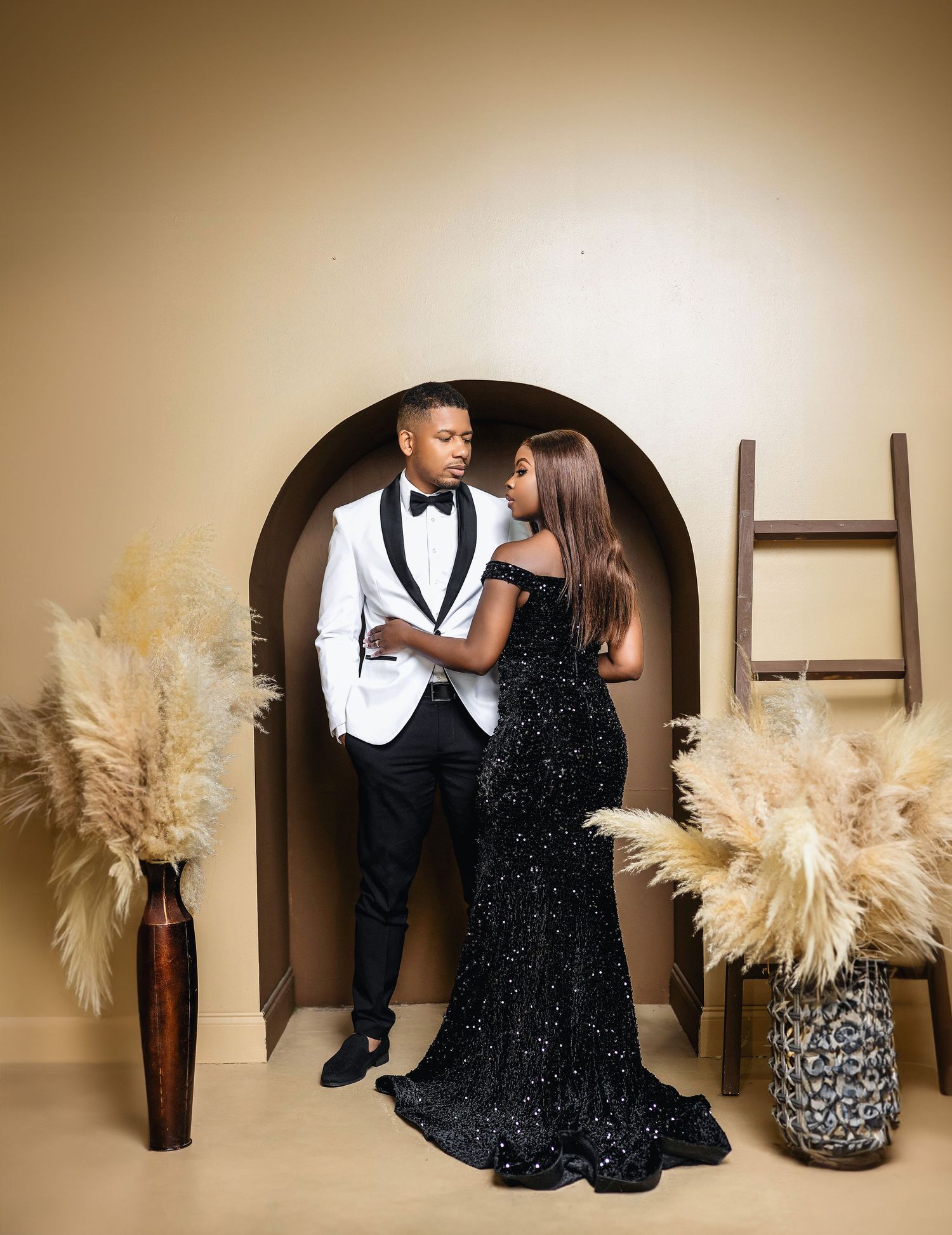 Elegant couple posing in arched alcove, man in white tuxedo and woman in black sequin gown with pampas grass decor.