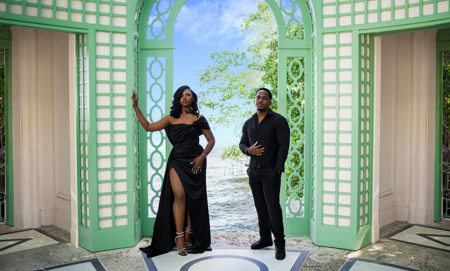 Couple in elegant black outfits posing in green lattice doorway with ocean view.