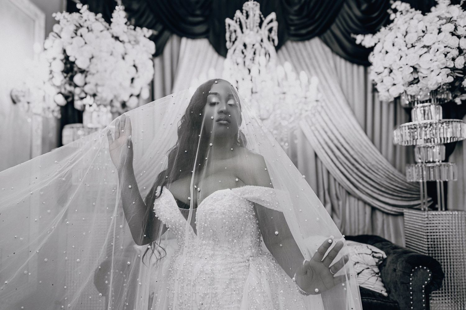 Black and white photo of a bride in a strapless gown and flowing veil, posing elegantly in a decorated wedding venue.