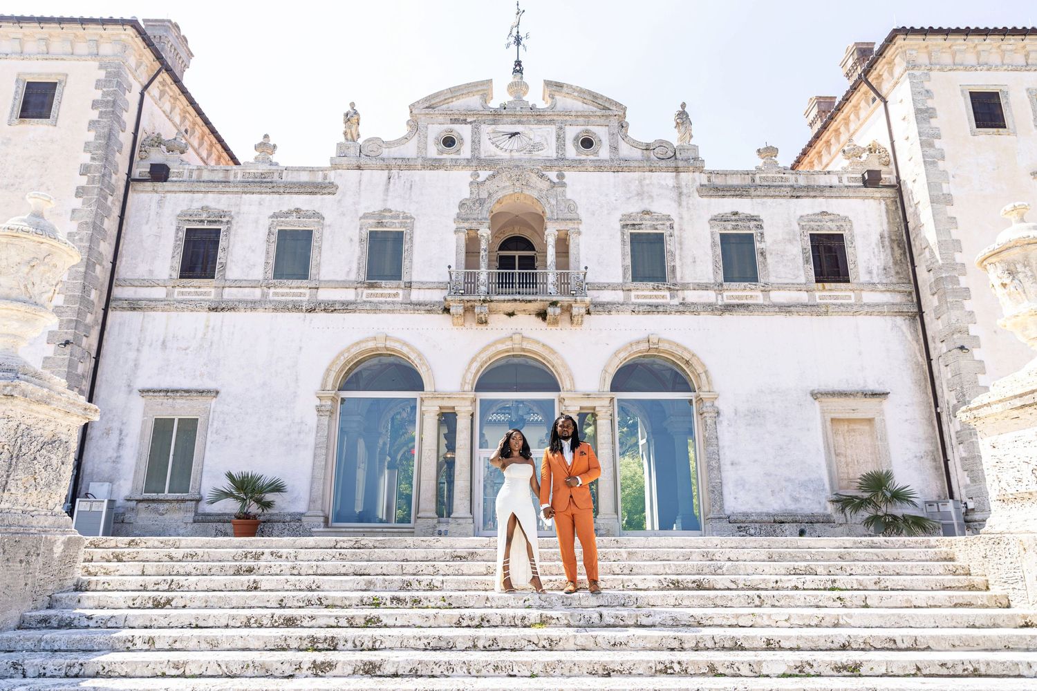 Couple in white gown and orange suit posing on grand steps of historic Mediterranean-style villa.