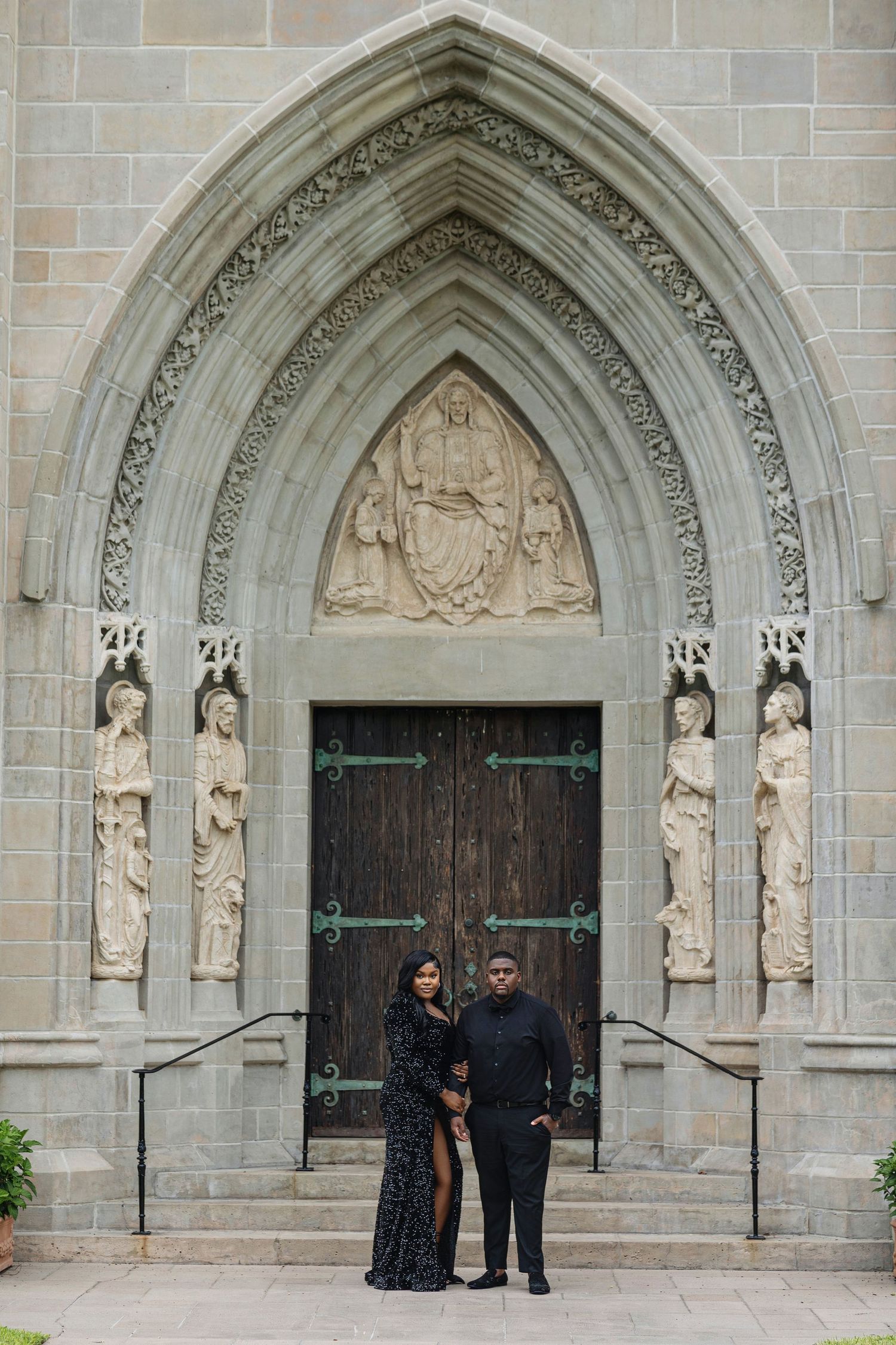 Couple in matching black formal attire embracing before grand Gothic cathedral doors with stone carvings.