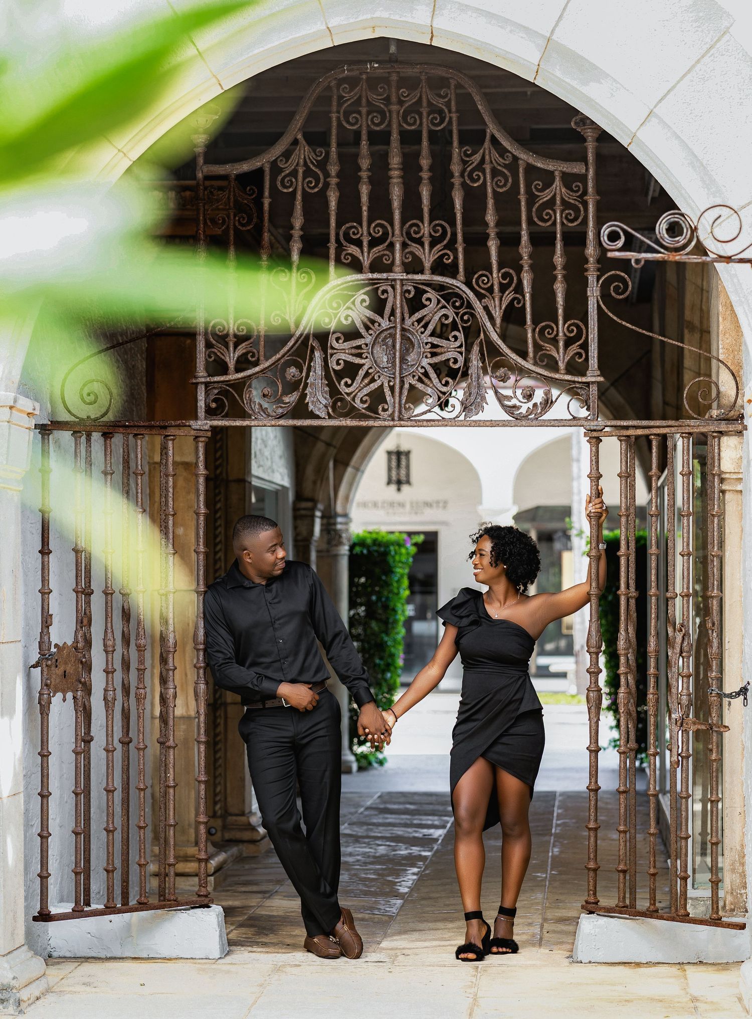 Couple in black outfits holding hands under an ornate wrought iron archway in a historic courtyard.