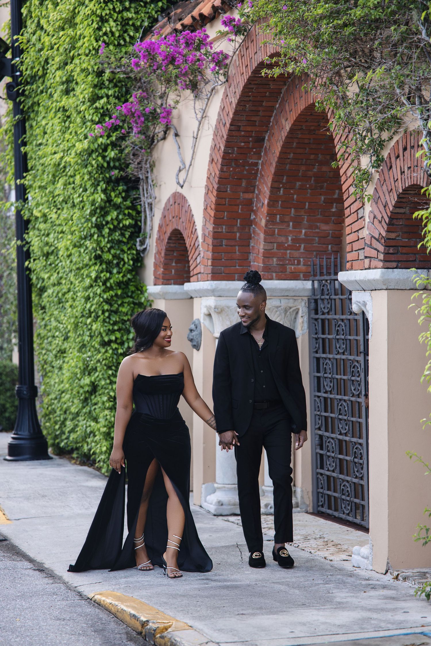 Stylish couple in black formal wear holding hands while strolling past ivy-covered arches.