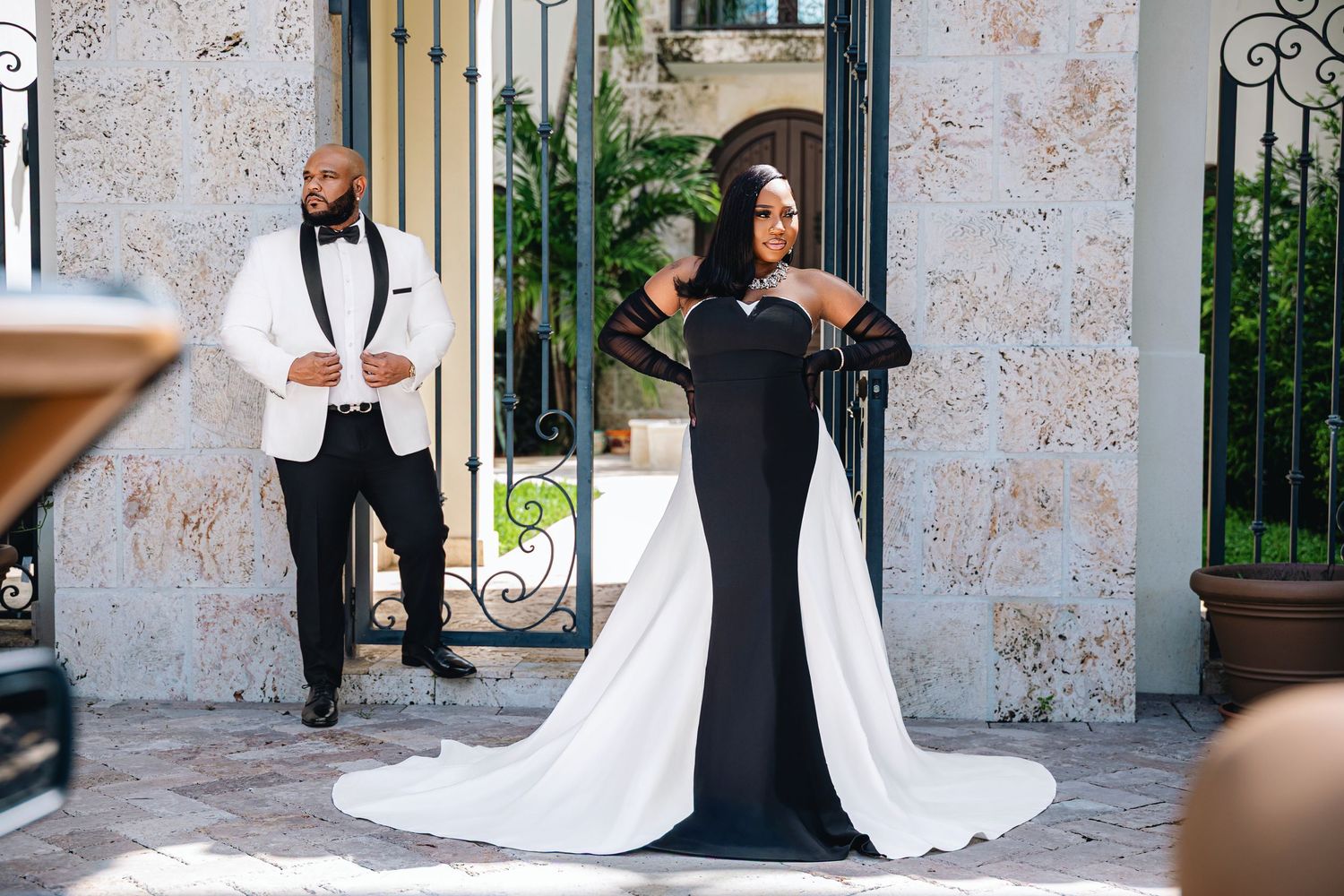 Bride in dramatic black and white gown with flowing train poses before ornate iron gates with groom in white tuxedo.