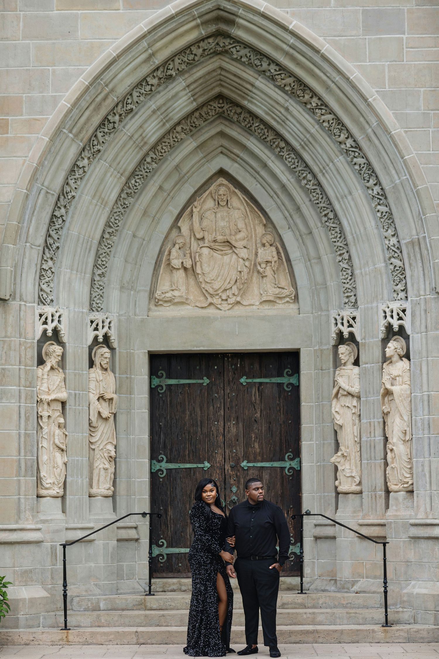 Couple in black formal attire standing before ornate Gothic cathedral doors with religious stone sculptures.
