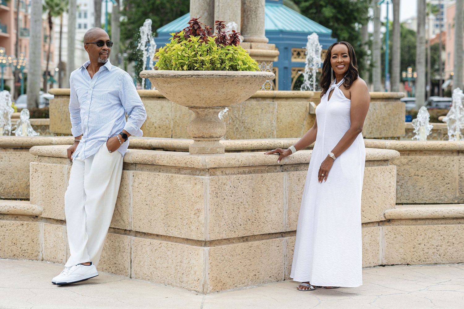 Couple in white attire posing on opposite sides of an elegant stone fountain outdoors.