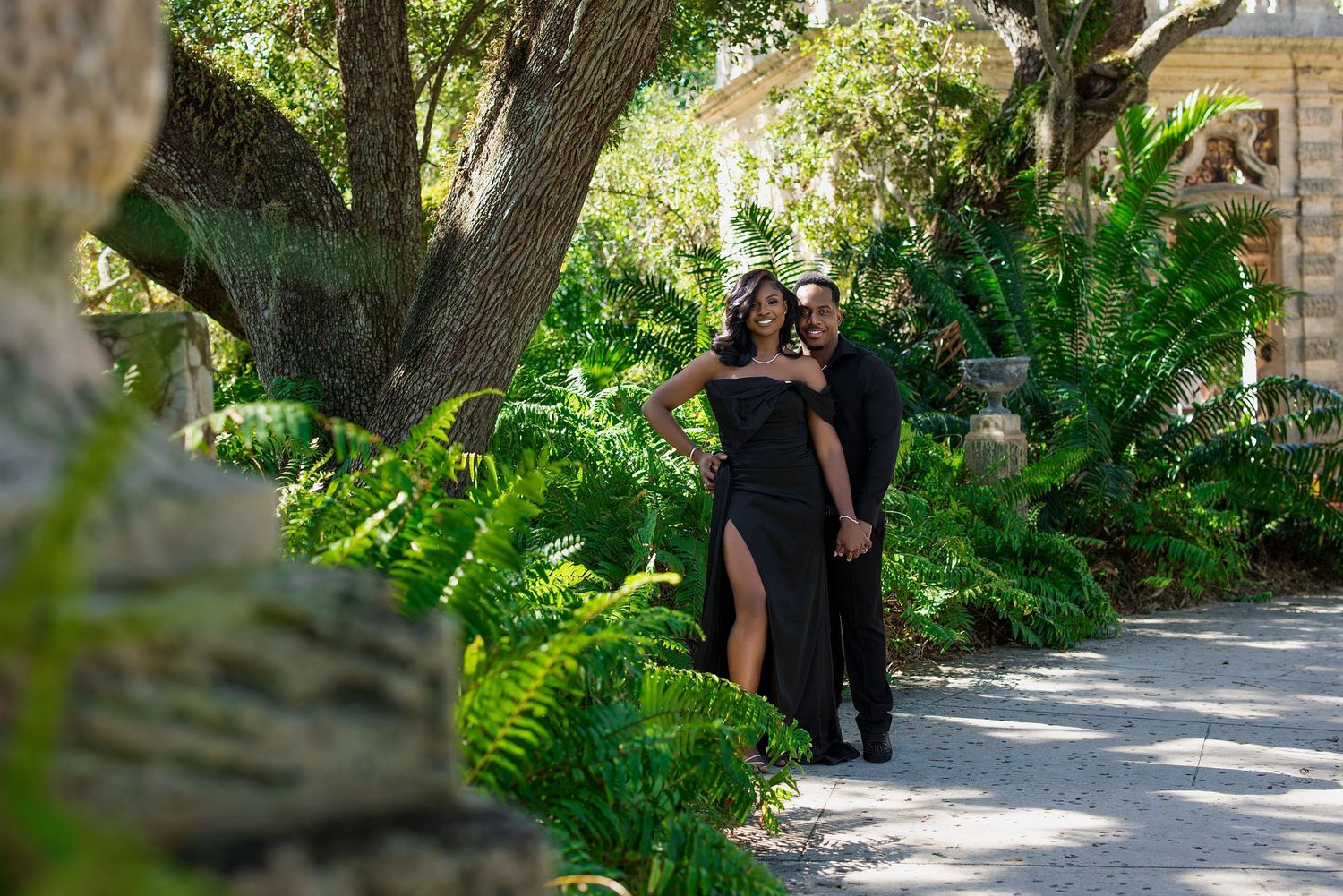 Elegant couple in black attire walking through a lush tropical garden path.
