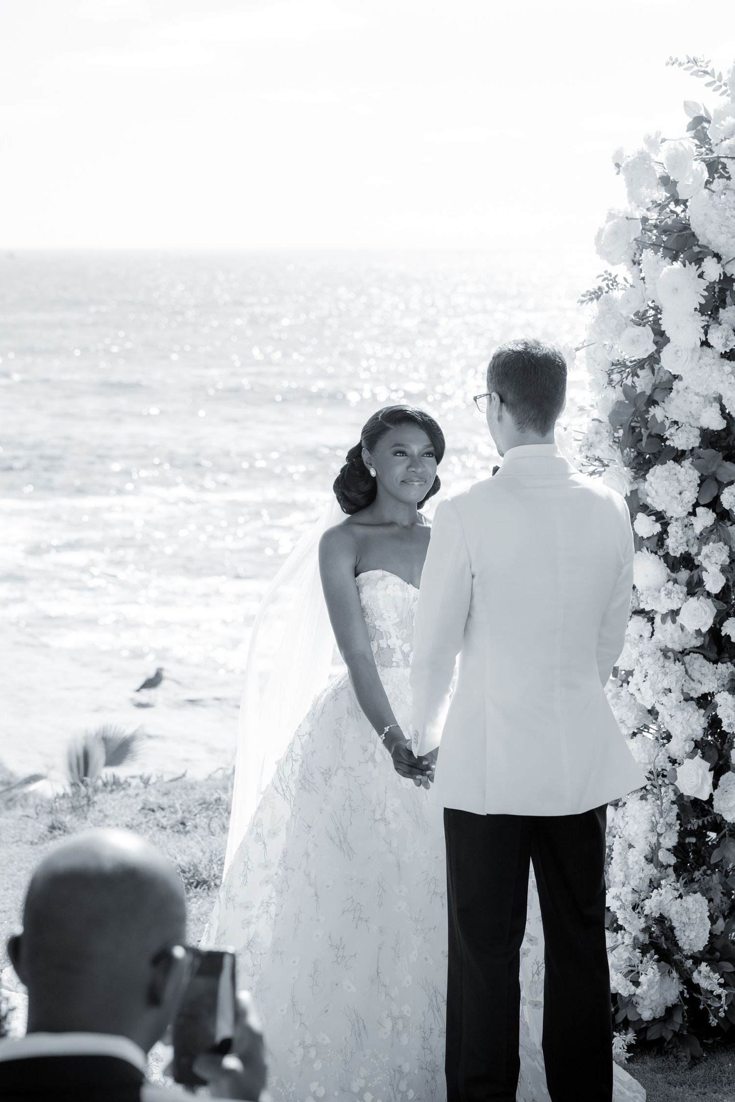 Black and white photo of a bride and groom exchanging vows by the ocean, flanked by a white floral arch.