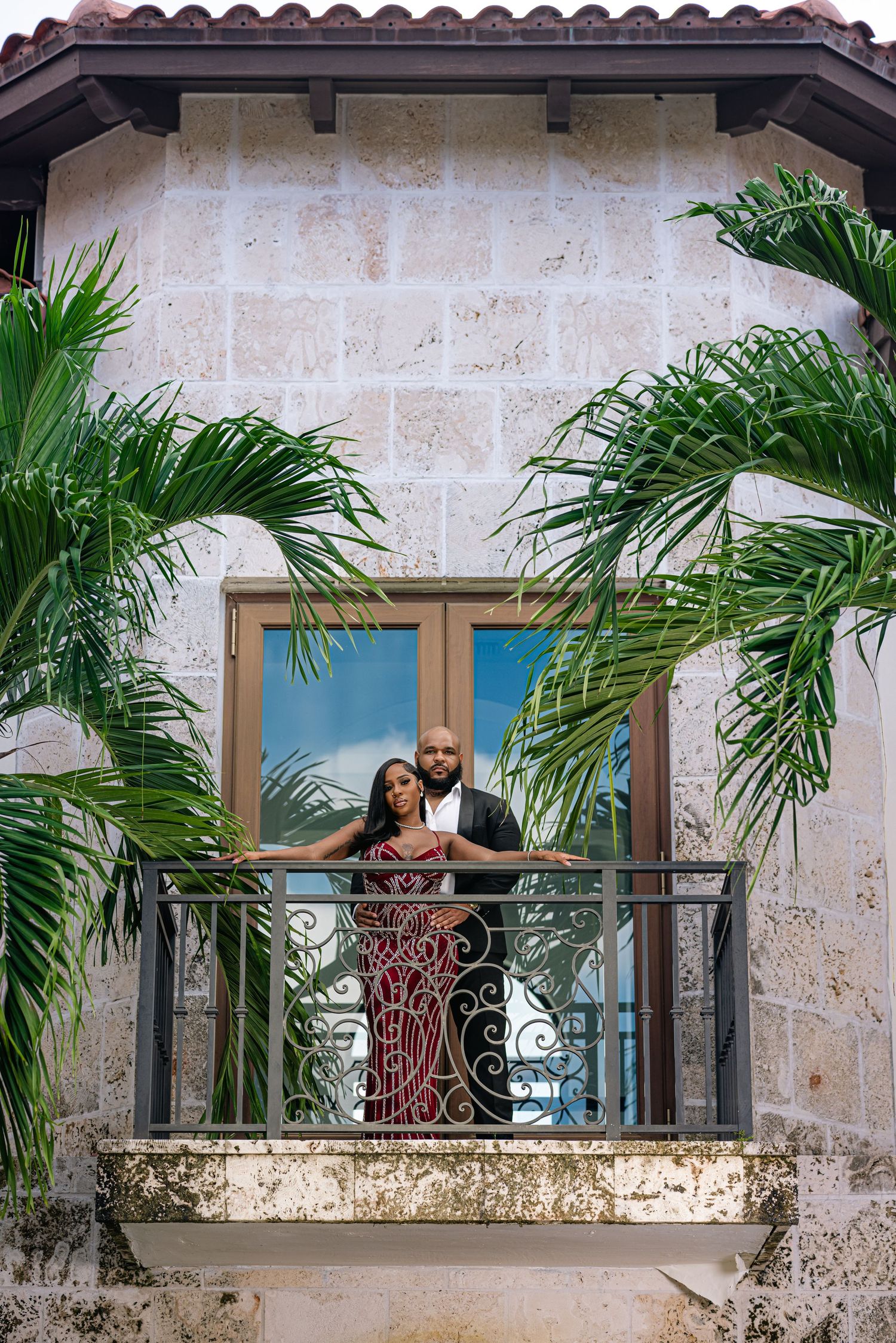 Couple stands on ornate balcony surrounded by tropical palm fronds at a luxurious stone estate.