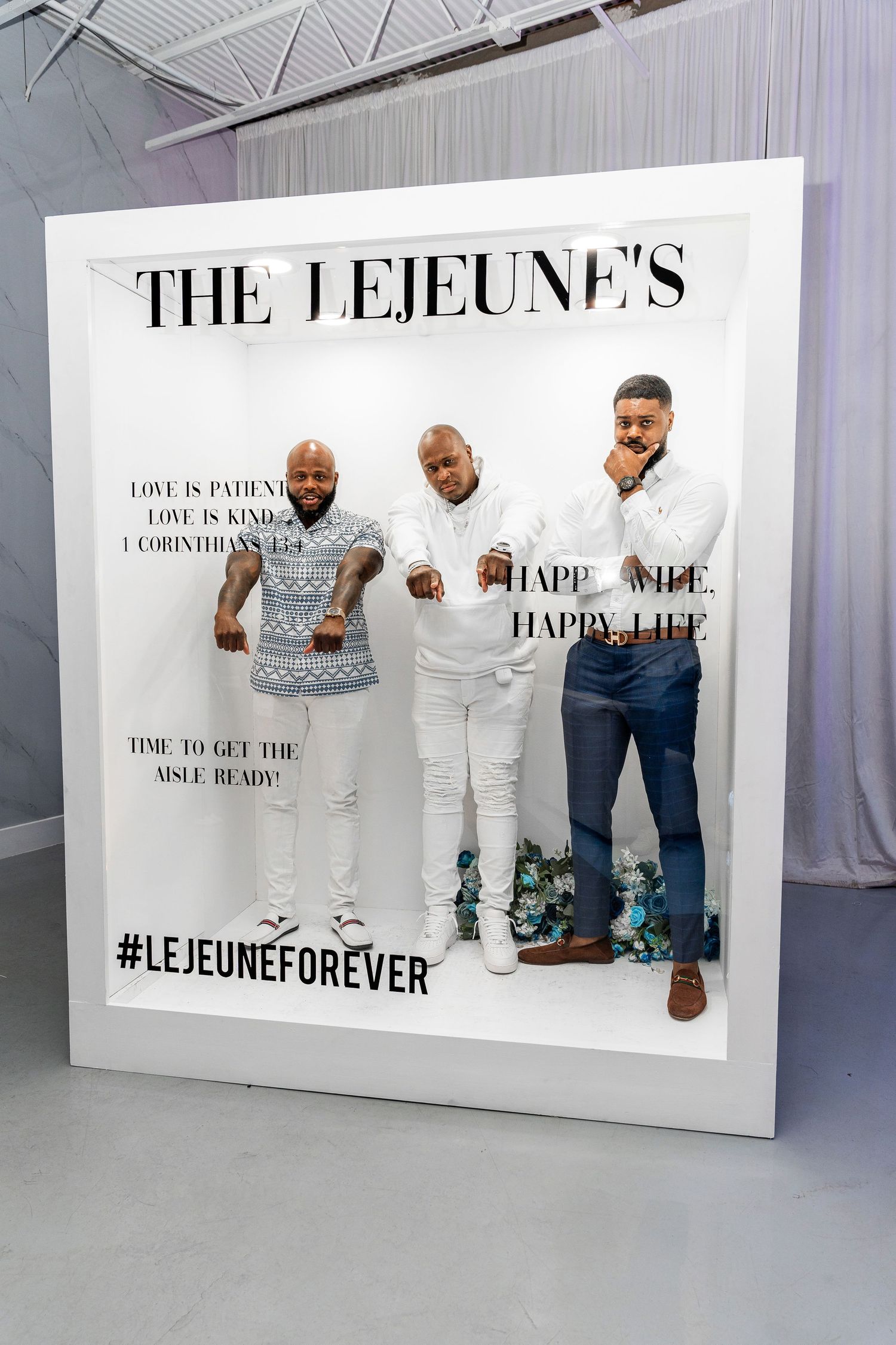 Three men in white outfits pose confidently inside The Lejeunes hashtag LejeuneForever photo frame backdrop.