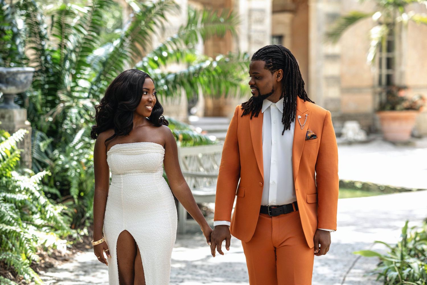Happy couple holding hands and smiling at each other in garden wearing white dress and orange suit.