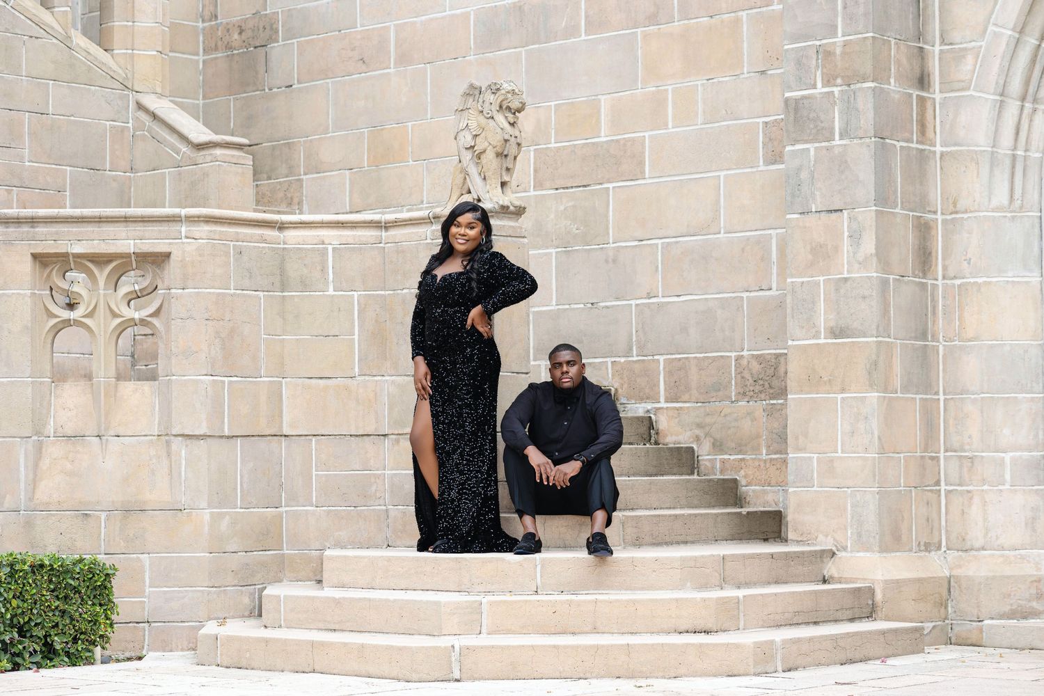 Elegant couple in all-black formal wear posing on stone steps outside a Gothic stone cathedral.