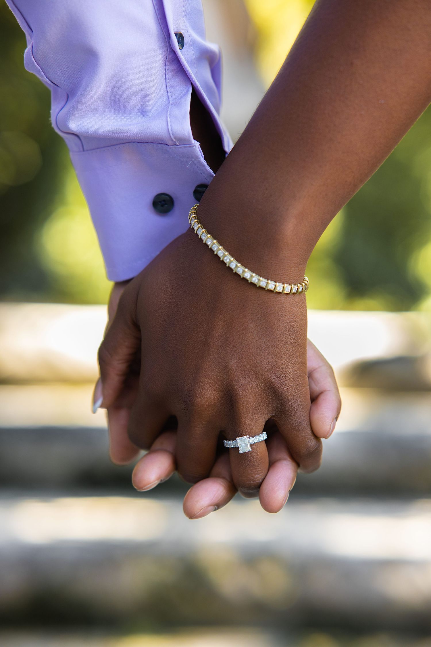 Close-up of Black couple holding hands, showcasing diamond engagement ring and tennis bracelet outdoors.