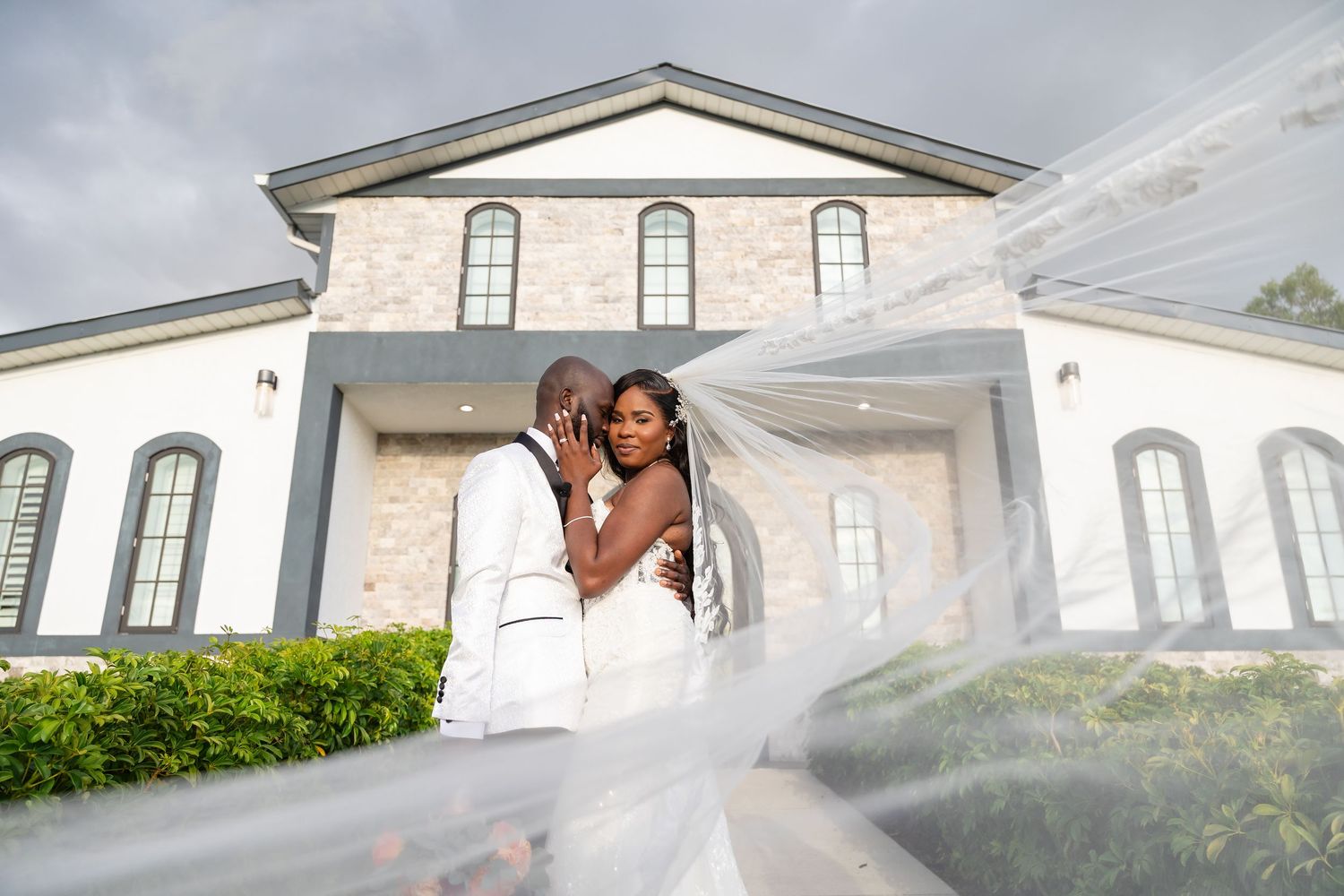 A bride and groom embrace in front of an elegant white church, her veil flowing dramatically in the wind.