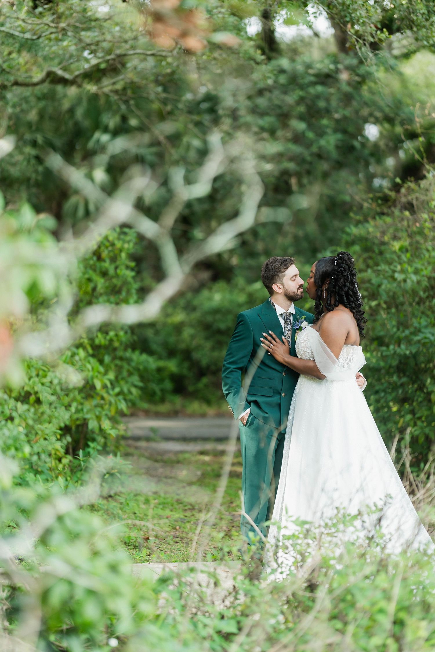 A bride and groom share a romantic moment surrounded by lush green trees in an outdoor wedding setting.