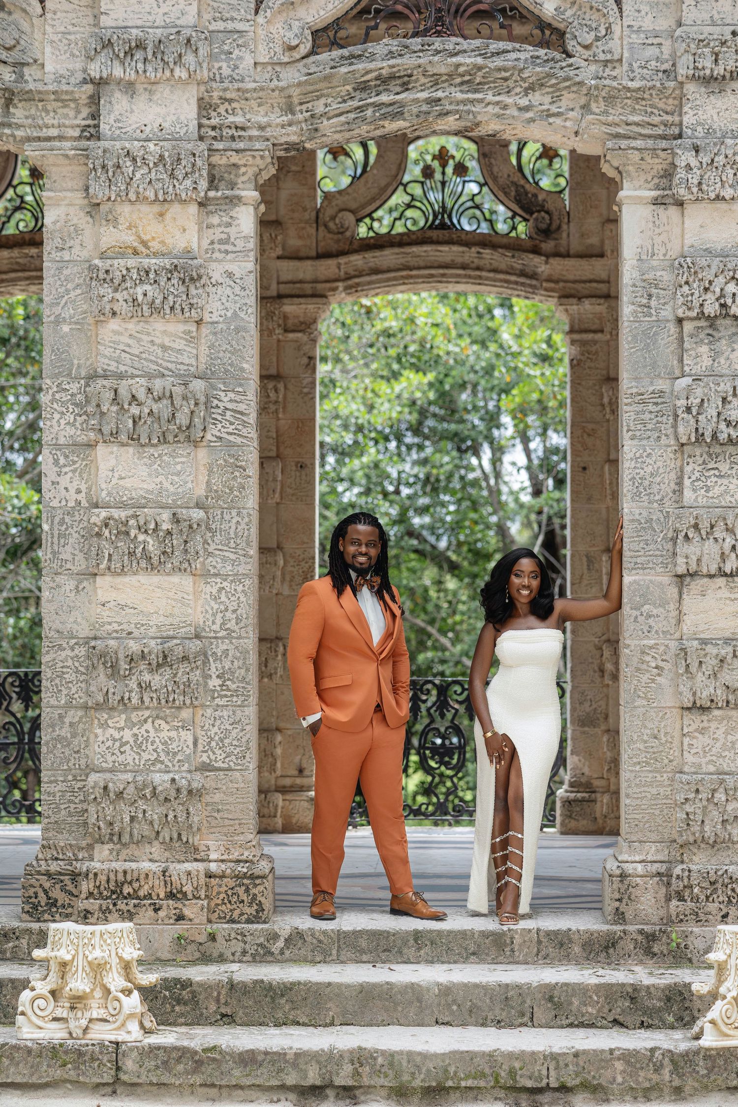 Couple posing under ornate carved stone archway, man in orange suit, woman in white dress.