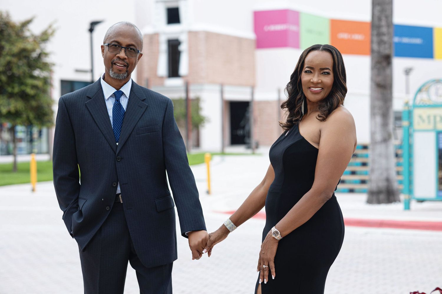 Engaged couple holding hands and smiling outdoors near colorful urban storefronts in coordinated formal attire.