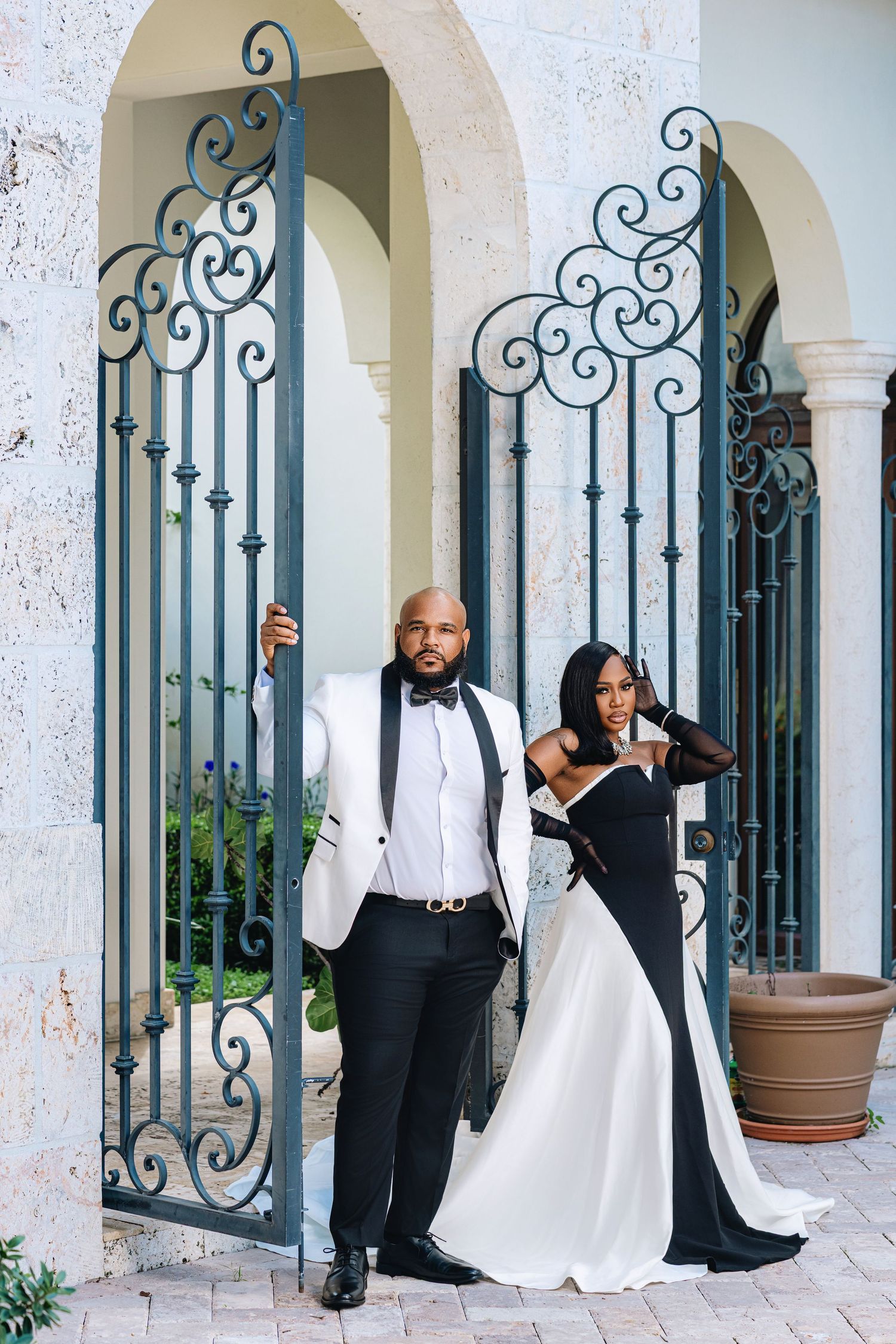 Couple in elegant black and white formalwear pose against decorative wrought iron gates at a grand estate.