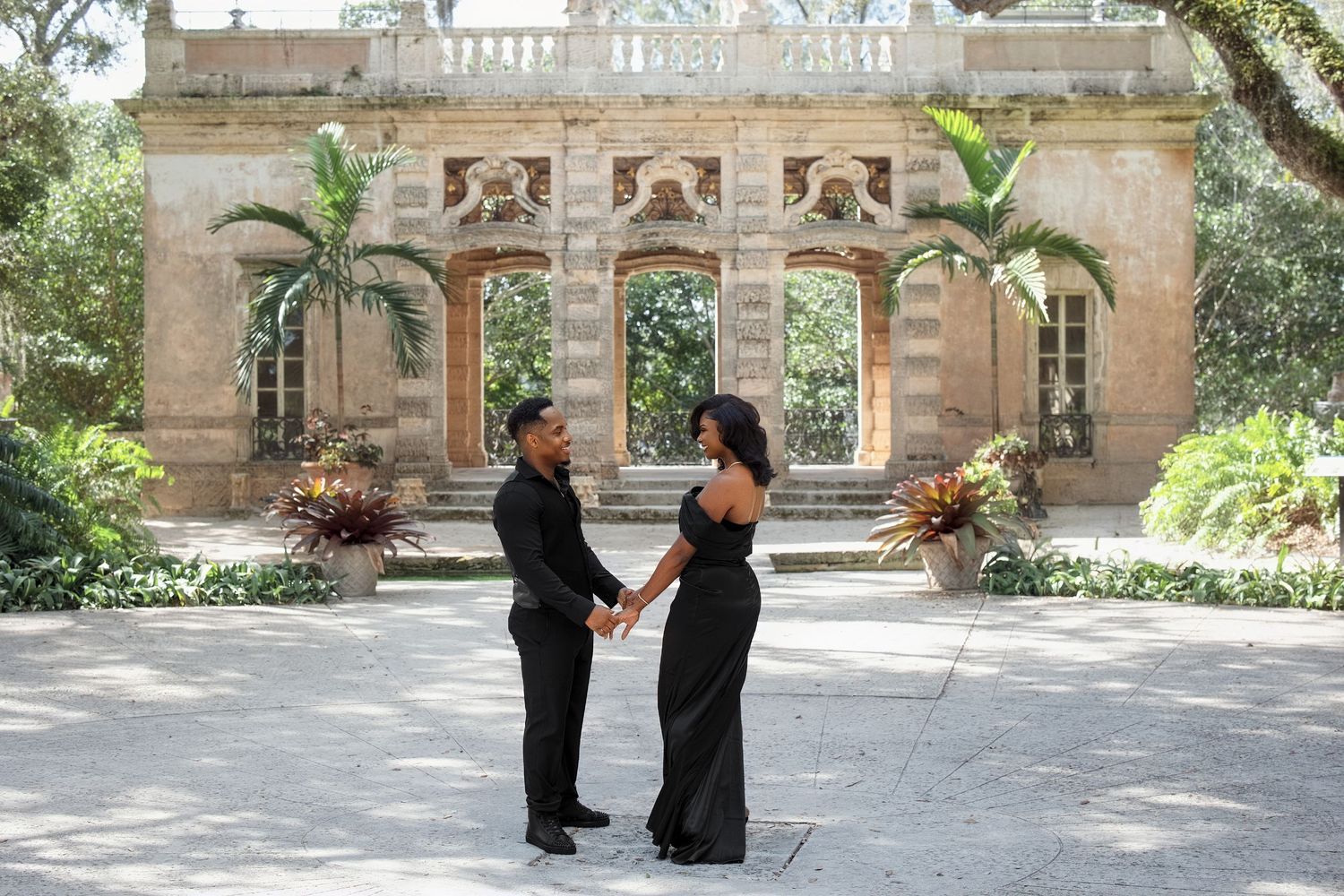 Black couple holding hands in matching black formal wear before ornate stone building in garden.