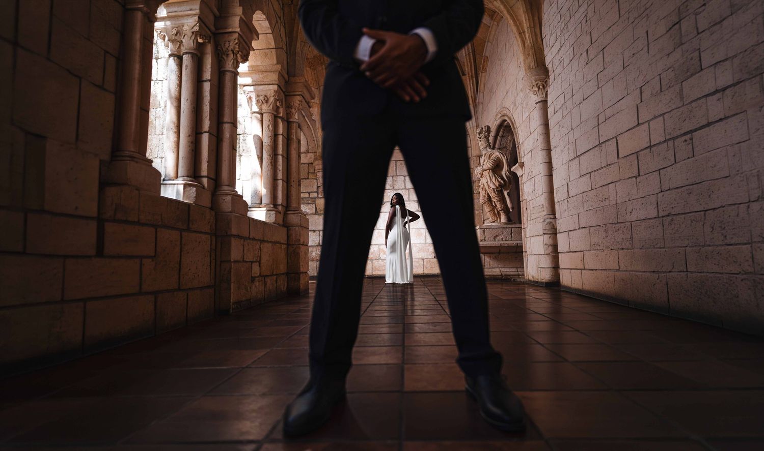 Man in dark suit stands in stone corridor with woman in white dress visible in background.