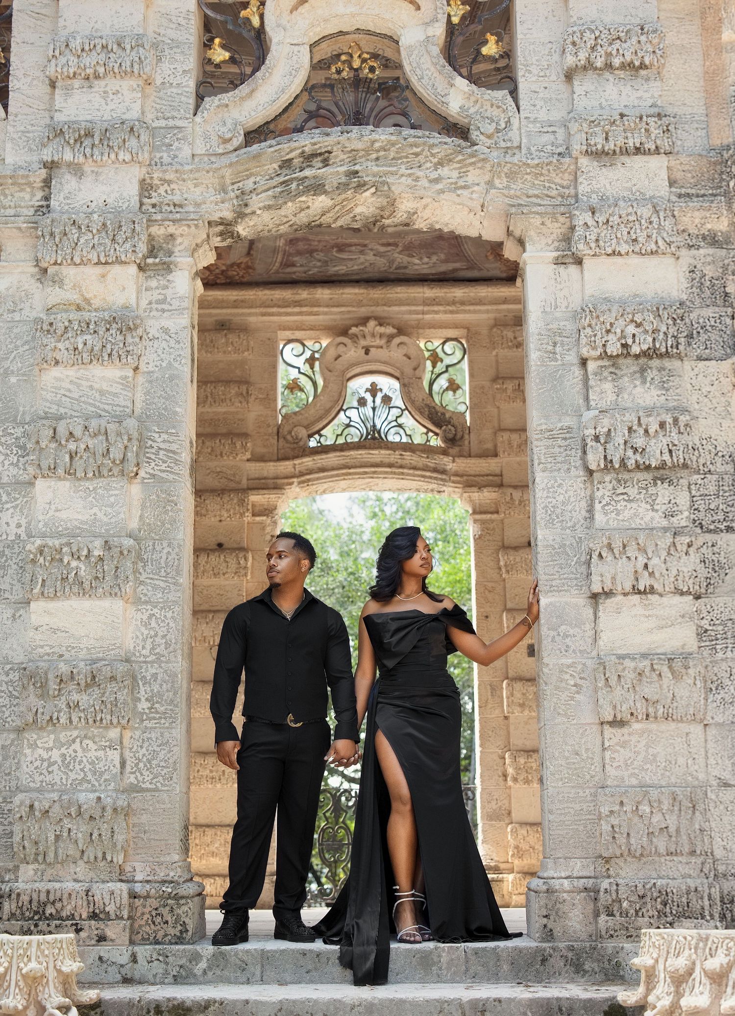 Couple in all-black outfits posing in ornate carved stone archway entrance.