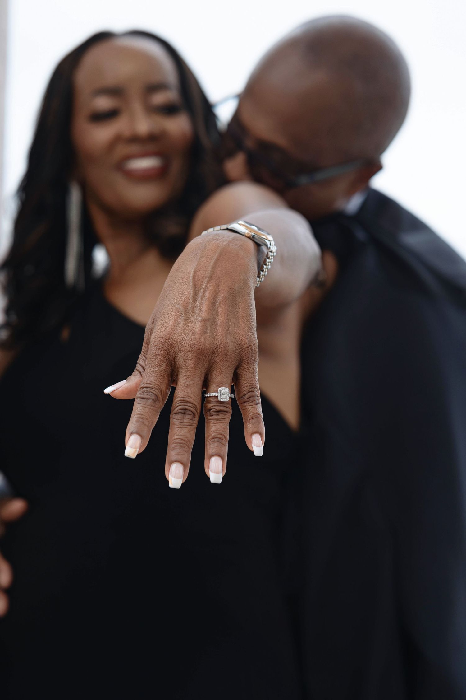 Smiling woman displays diamond engagement ring while partner kisses her cheek in a joyful proposal moment.