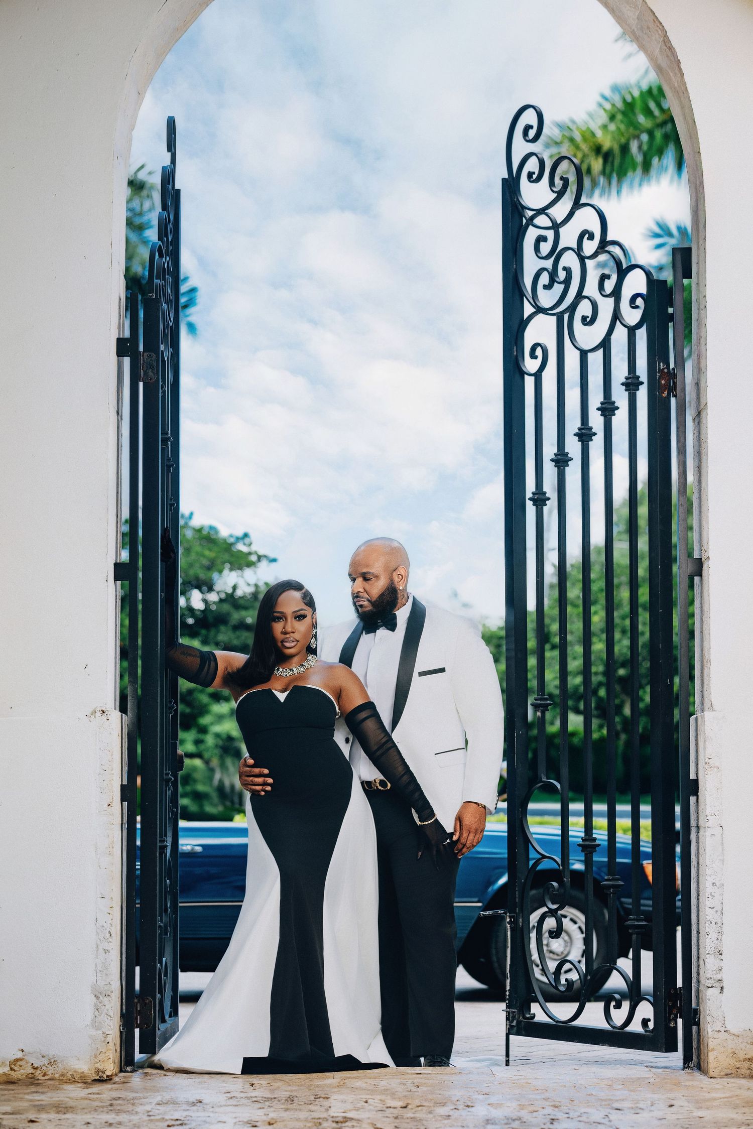 Elegant Black couple in black and white formal attire pose under ornate iron gate archway.