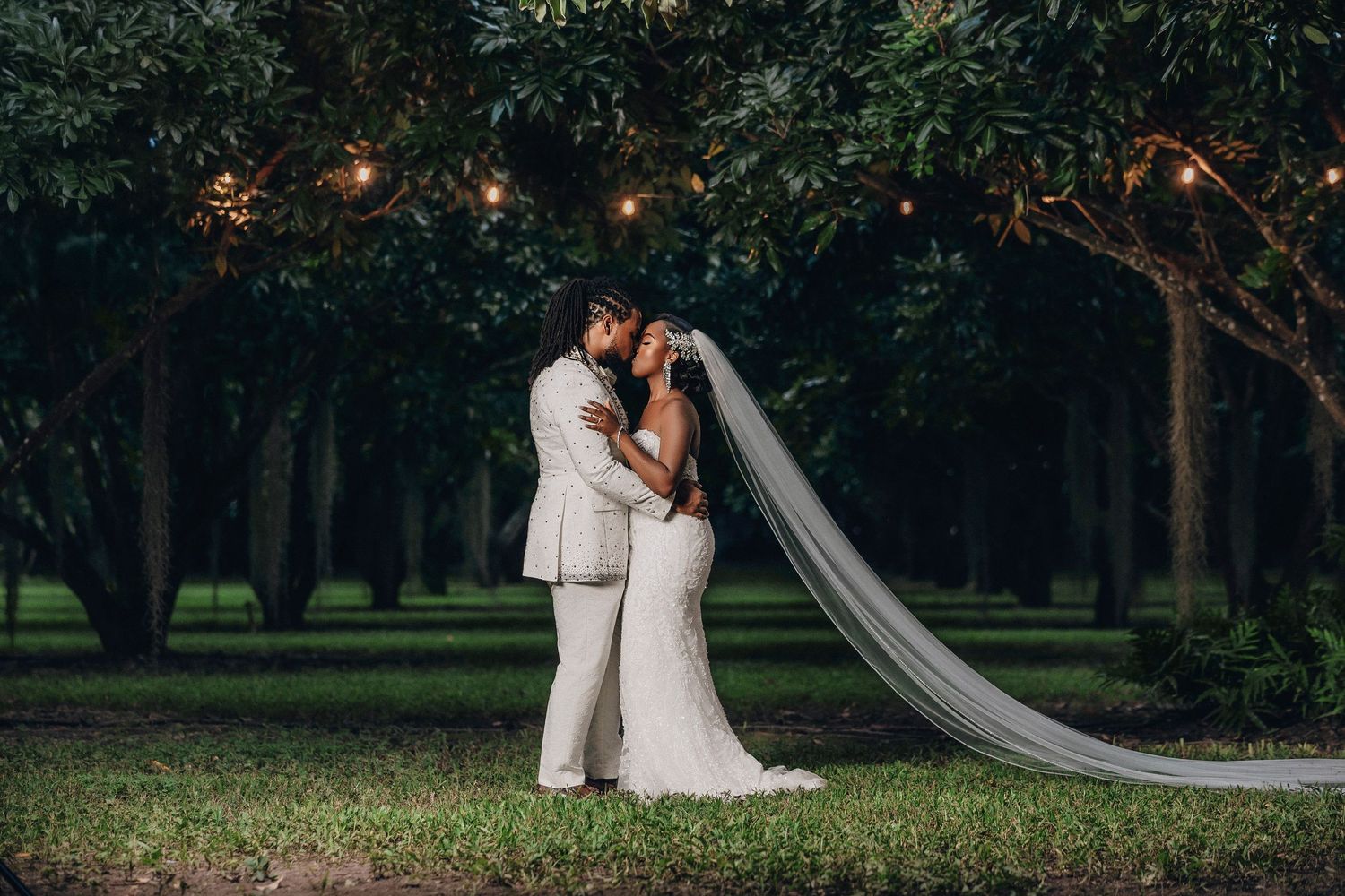 Bride and groom share a romantic embrace under string lights in a lush green garden at dusk, wedding veil flowing.