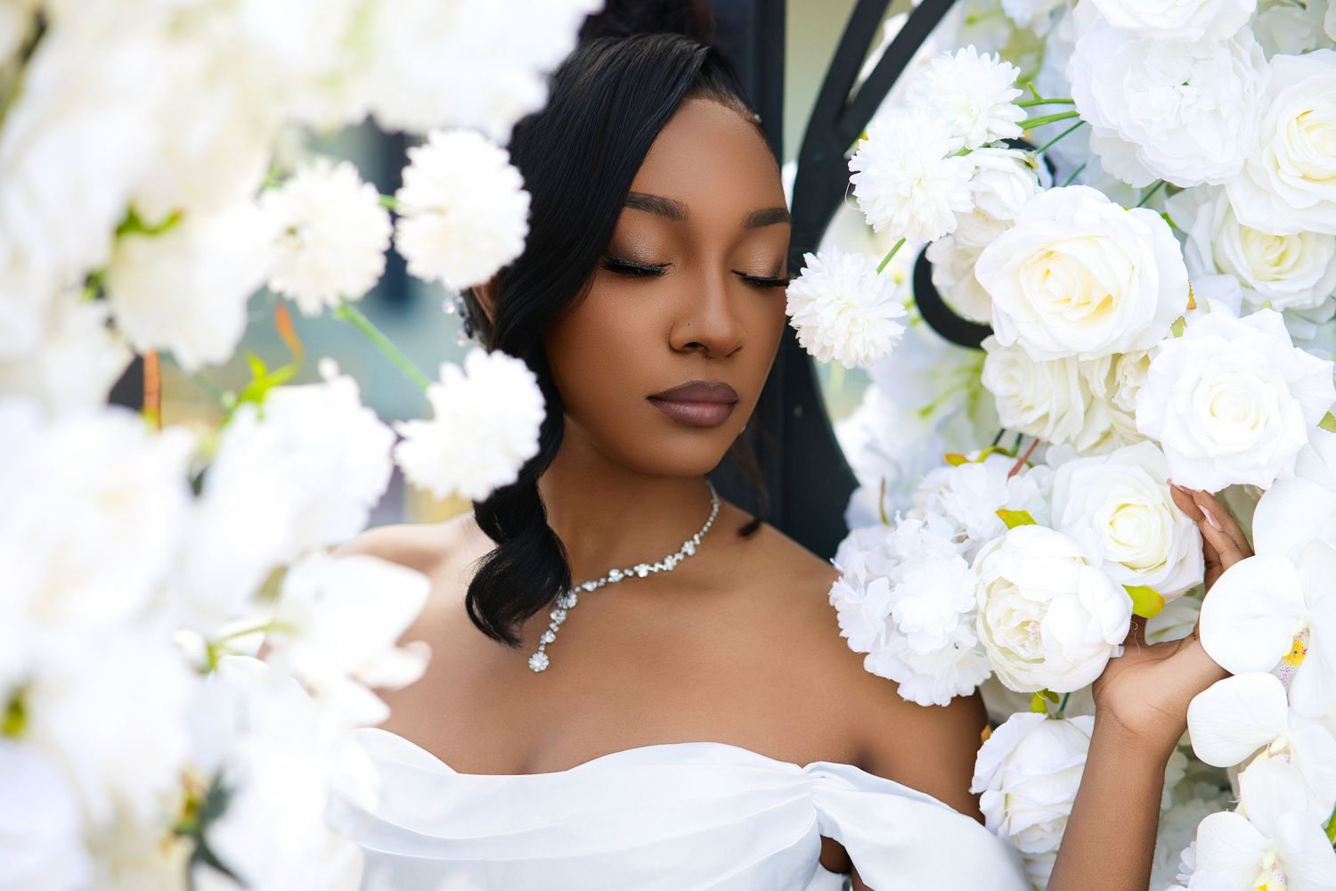Elegant bride in white strapless gown wearing a diamond necklace surrounded by lush white roses.
